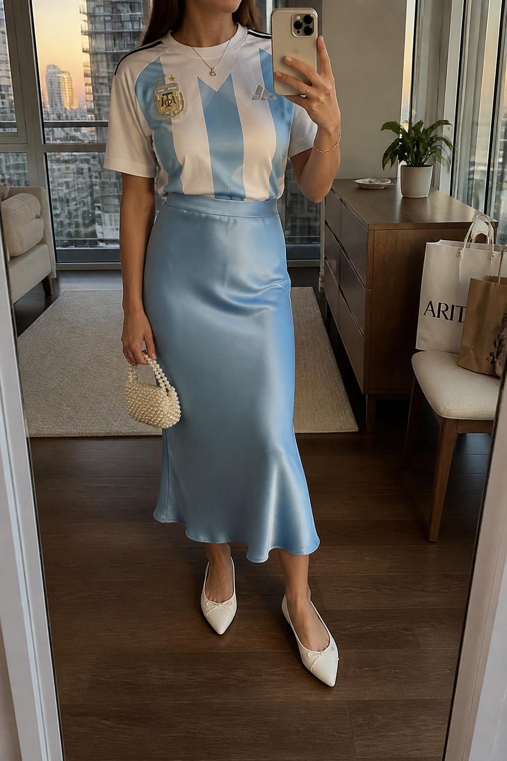 mirror selfie of a woman wearing a white Argentina soccer jersey tucked into a powder blue satin midi skirt, white pointed ballet flats, pearl mini handbag, and delicate silver necklace, clearly showing the same clothing items, colors, fabrics, and accessories listed in the Outfit Pieces section, holding a phone in front of a mirror, cropped at NECK so the face does not show, with a relaxed casual selfie pose, in a stylish city apartment, evening golden hour, including a small plant on a dresser and shopping bags beside a chair, latest iPhone photo quality.