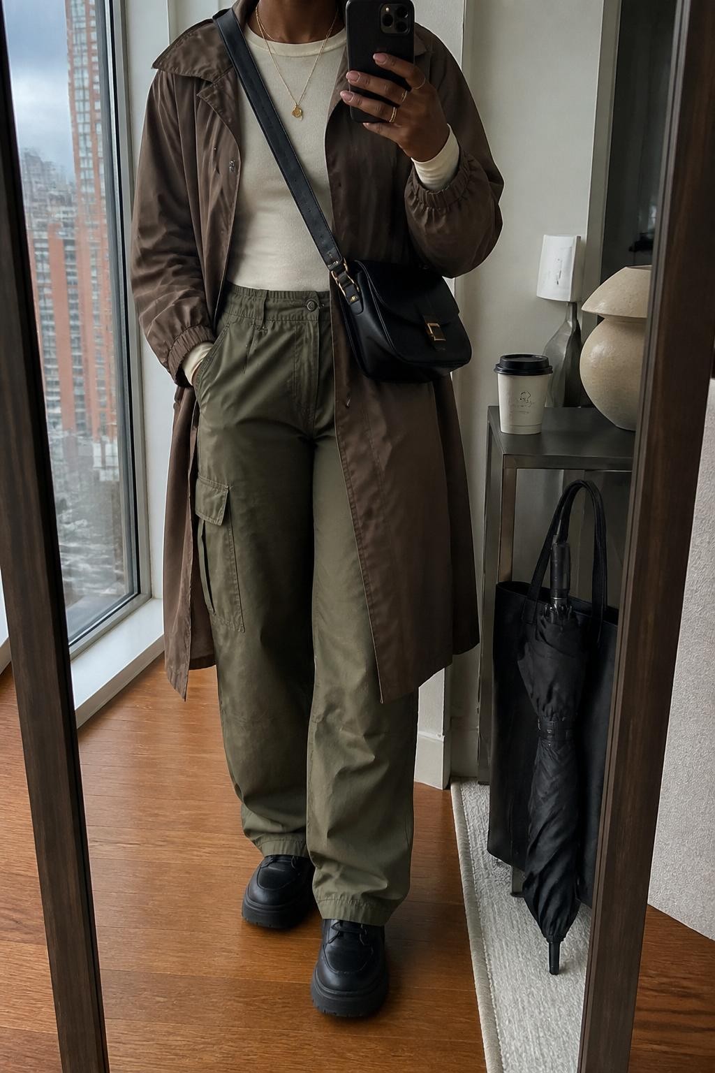 mirror selfie of a Black woman wearing the exact outfit described above, clearly showing a mocha waterproof trench coat layered over a fitted cream long-sleeve top, olive cargo pants, black waterproof platform sneakers, a black crossbody bag, and gold layered necklaces, holding a phone in front of a mirror, cropped at NECK so the face does not show, with a relaxed casual selfie pose, in a stylish city apartment mirror, gray afternoon light, with a compact umbrella beside a tote bag and a coffee cup on a side table, latest iPhone photo quality.