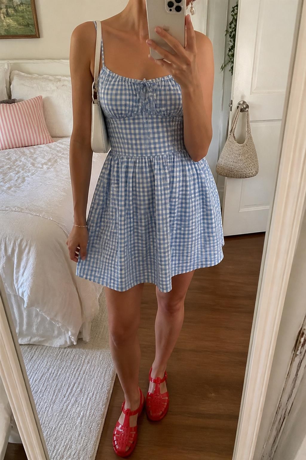 mirror selfie of a woman wearing a blue and white gingham sundress, glossy red jelly shoes, white mini shoulder bag, pearl stud earrings, and a slim silver bracelet, clearly showing the same clothing items, colors, fabrics, and accessories listed in the Outfit Pieces section, holding a phone in front of a mirror, cropped at NECK so the face does not show, with a relaxed casual selfie pose, in a cozy bedroom mirror setup, natural window light, including soft pillows on an unmade bed and a handbag hanging on a door handle, latest iPhone photo quality.