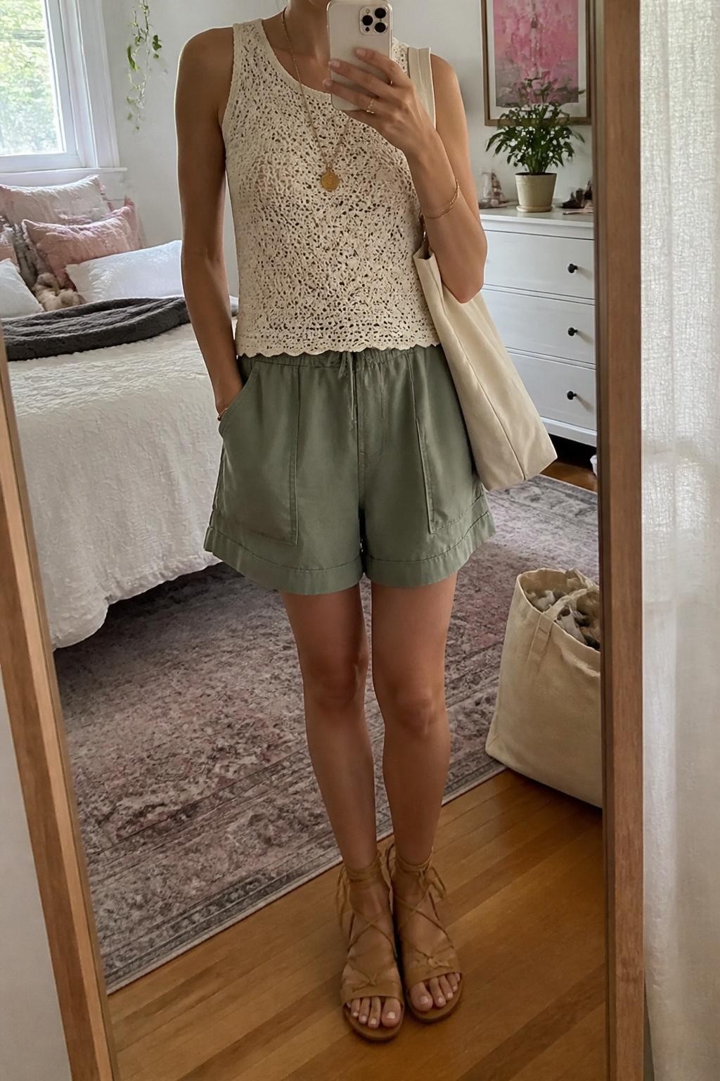 mirror selfie of a woman wearing sage green baggy cargo shorts, ivory sleeveless crochet top, tan lace-up flat sandals, cream canvas tote bag, and gold charm necklace, clearly showing the same clothing items, colors, fabrics, and accessories listed in the Outfit Pieces section, holding a phone in front of a mirror, cropped at NECK so the face does not show, with a relaxed casual selfie pose, in a feminine-themed bedroom, natural window light, including a cream tote bag resting on the floor and a small plant on a dresser, latest iPhone photo quality.