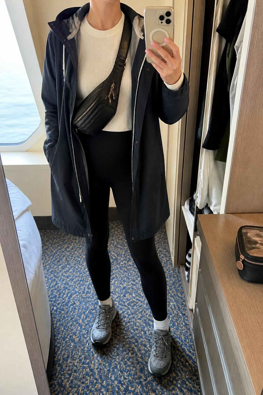 Outfit Image Prompt:
mirror selfie of a woman wearing a navy hooded raincoat, white long-sleeve thermal top, black high-waisted leggings, gray waterproof hiking sneakers, a black belt bag worn crossbody, and simple silver studs, holding a phone in front of a mirror, cropped at NECK so the face does not show, relaxed casual selfie pose, in a minimal cruise cabin mirror corner with natural window light, including an open closet with clothes visible and a small makeup bag on the counter, realistic latest iPhone photo quality, natural casual mirror selfie.