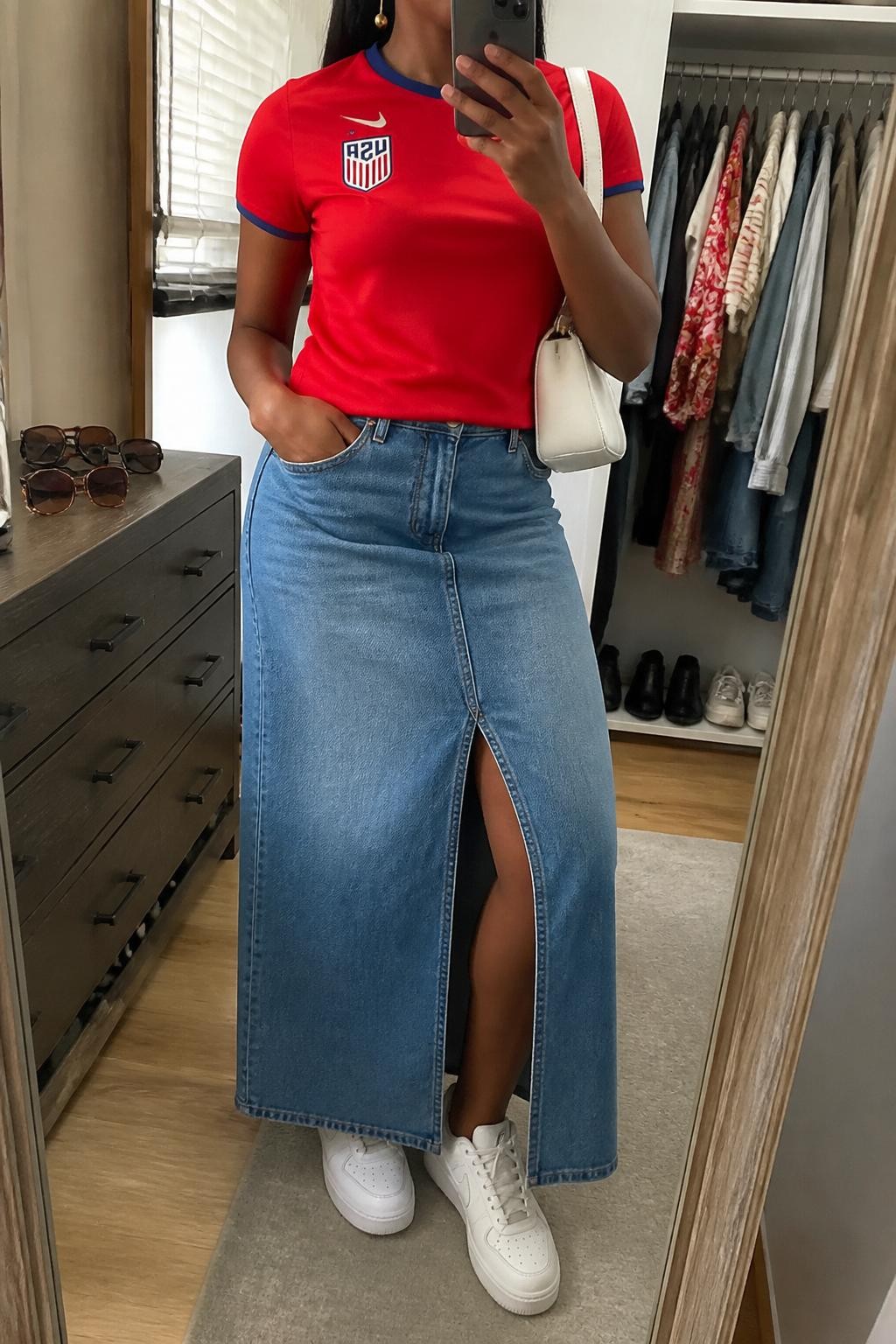 mirror selfie of a Black woman wearing a red USA soccer jersey, medium blue denim maxi skirt with front slit, white leather sneakers, white crescent shoulder bag, and gold drop earrings, clearly showing the same clothing items, colors, fabrics, and accessories listed in the Outfit Pieces section, holding a phone in front of a mirror, cropped at NECK so the face does not show, with a relaxed casual selfie pose, in a walk-in closet mirror, natural window light, including an open closet with clothes visible and sunglasses placed on a dresser, latest iPhone photo quality.