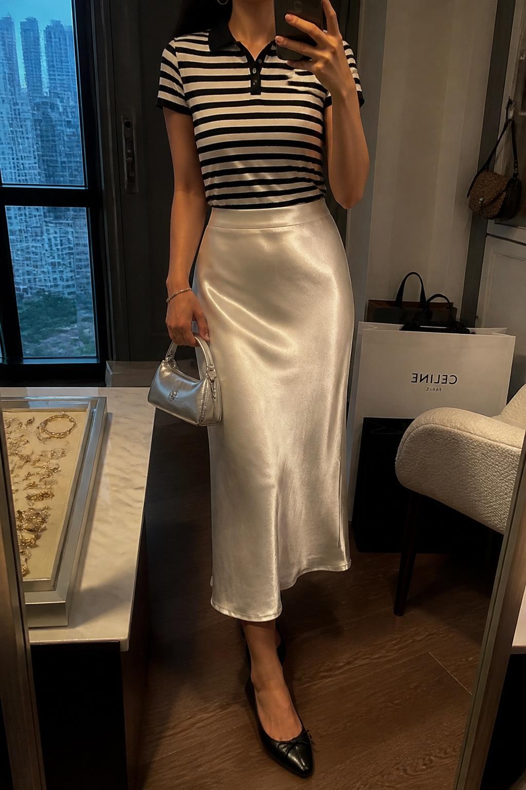 mirror selfie of a woman wearing a black and white striped fitted polo shirt tucked into an ivory satin midi skirt, black pointed ballet flats, silver mini handbag, and delicate silver hoop earrings, clearly showing the same clothing items, colors, fabrics, and accessories listed in the Outfit Pieces section, holding a phone in front of a mirror, cropped at NECK so the face does not show, with a relaxed casual selfie pose, in a stylish city apartment, warm moody lighting, including a jewelry tray on the vanity and shopping bags beside a chair, latest iPhone photo quality.