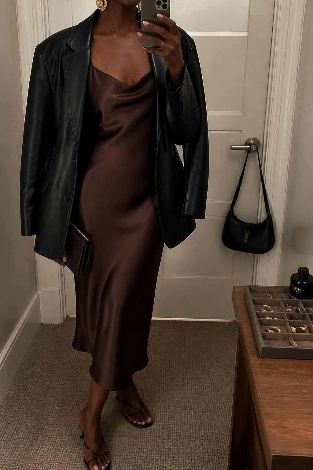 Outfit Image Prompt:
mirror selfie of a Black woman with deep brown skin wearing a chocolate brown satin slip dress, oversized black leather blazer, black strappy kitten heels, small chocolate brown clutch bag, and gold statement earrings, holding a phone in front of a mirror, cropped at NECK so the face does not show, relaxed high-fashion mirror selfie pose, in a minimal dressing room, warm moody lighting, with a jewelry tray on the vanity and a handbag hanging on a door handle, natural skin texture, realistic latest iPhone photo quality.