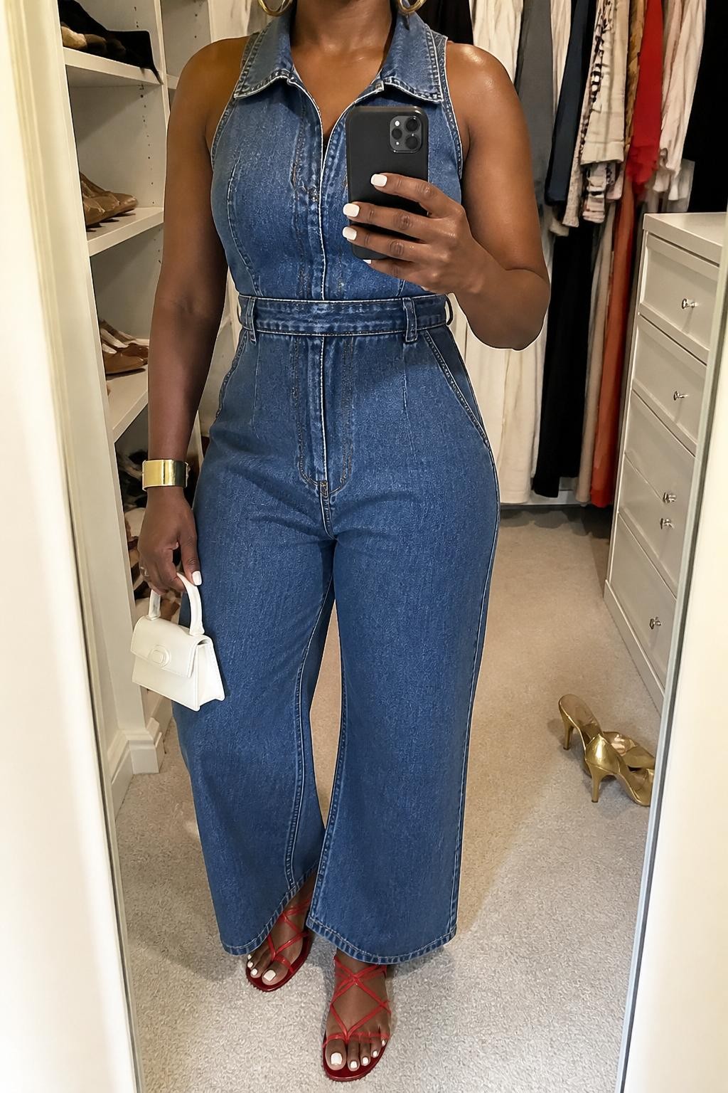 Outfit Image Prompt:
mirror selfie of a Black woman with rich brown skin wearing a blue denim sleeveless jumpsuit, red strappy flat sandals, a white mini handbag, gold cuff bracelet, and gold hoop earrings, clearly showing the exact outfit pieces, holding a phone in front of a mirror, cropped at NECK so the face does not show, relaxed casual selfie pose, in a walk-in closet mirror, soft indoor lighting, with an open closet with clothes visible and a pair of heels on the floor nearby, realistic latest iPhone photo quality, natural casual mirror selfie.