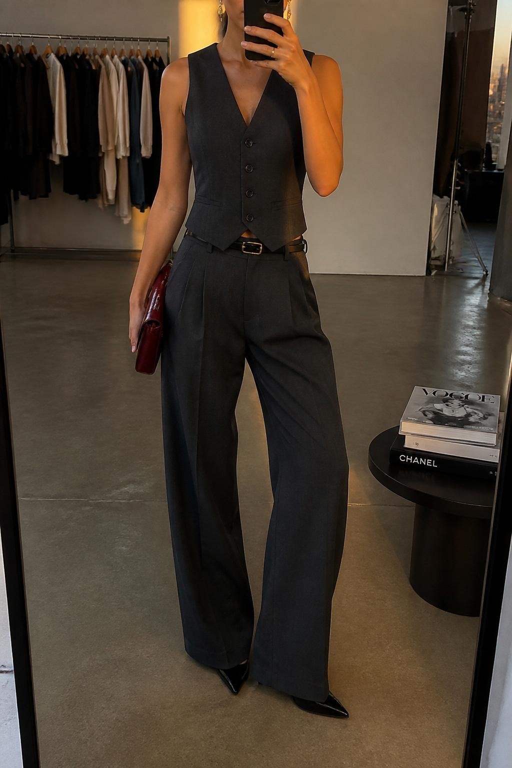 Outfit Image Prompt:
mirror selfie of a woman wearing a charcoal gray tailored waistcoat, matching charcoal wide-leg trousers, black pointed pumps, small burgundy clutch bag, gold drop earrings, and a slim black belt, holding a phone in front of a mirror, cropped at NECK so the face does not show, relaxed polished selfie pose, in a fashion studio mirror corner, evening golden hour lighting, with a clothing rack with garments behind and a stack of fashion magazines on a side table, realistic latest iPhone photo quality, NYC high fashion outfit.