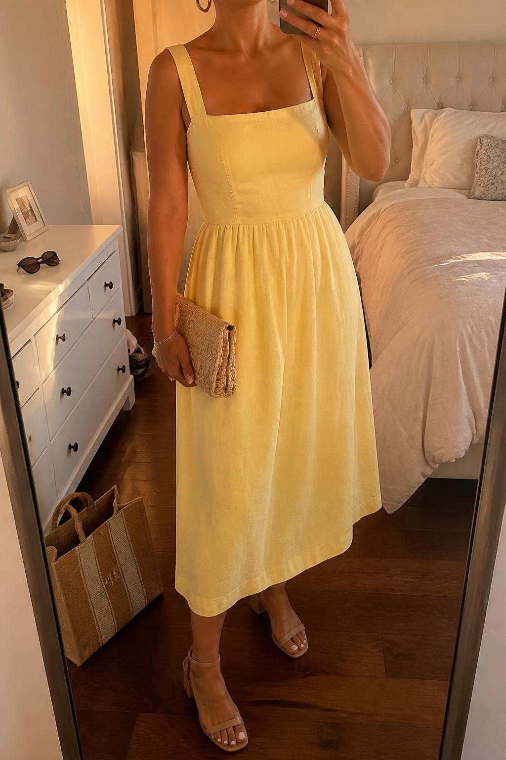 mirror selfie of a woman wearing a soft lemon linen midi dress with nude strappy block heels, medium gold hoop earrings, a natural woven clutch, and hair in a low bun with soft face-framing pieces, holding a phone in front of a mirror, cropped at NECK so face doesn't show, with a natural casual selfie pose, in a feminine-themed bedroom, evening golden hour, including small lived-in details like a tote bag on the floor, sunglasses on a dresser, or soft bedding in the background, latest iPhone photo quality (no watermarks on images)