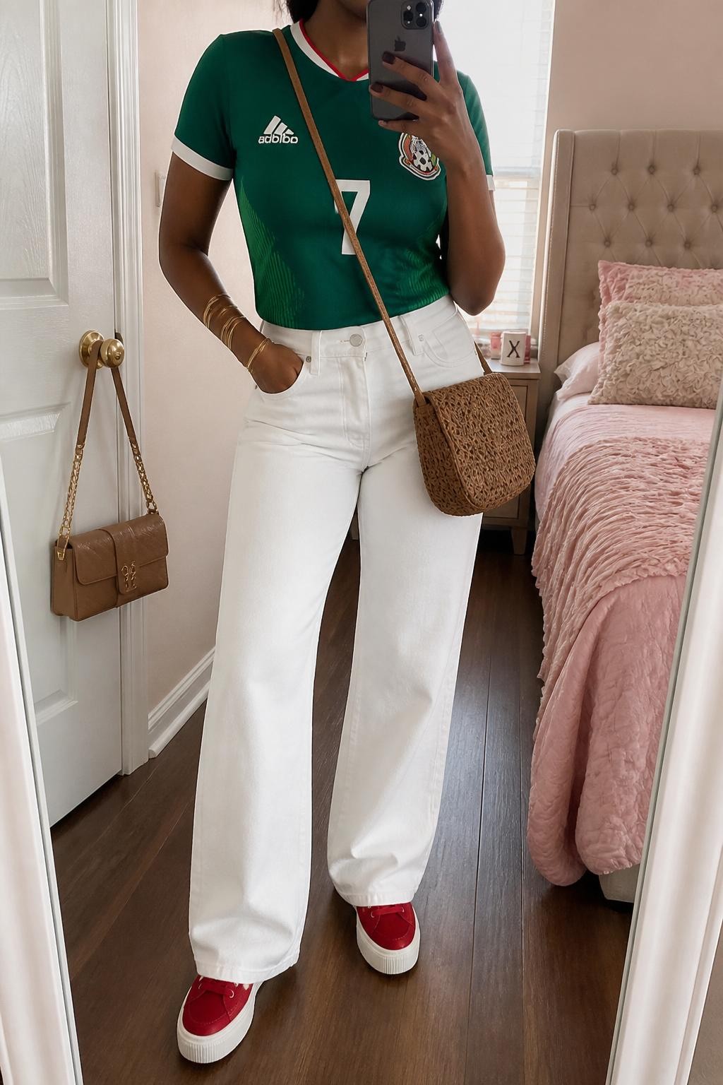 mirror selfie of a Black woman wearing a fitted green Mexico soccer jersey tucked into white high-waisted wide-leg jeans, red platform sneakers, tan woven crossbody bag, and gold bangle bracelet, clearly showing the same clothing items, colors, fabrics, and accessories listed in the Outfit Pieces section, holding a phone in front of a mirror, cropped at NECK so the face does not show, with a relaxed casual selfie pose, in a feminine-themed bedroom, natural window light, including a handbag hanging on a door handle and a coffee mug on a bedside table, latest iPhone photo quality.