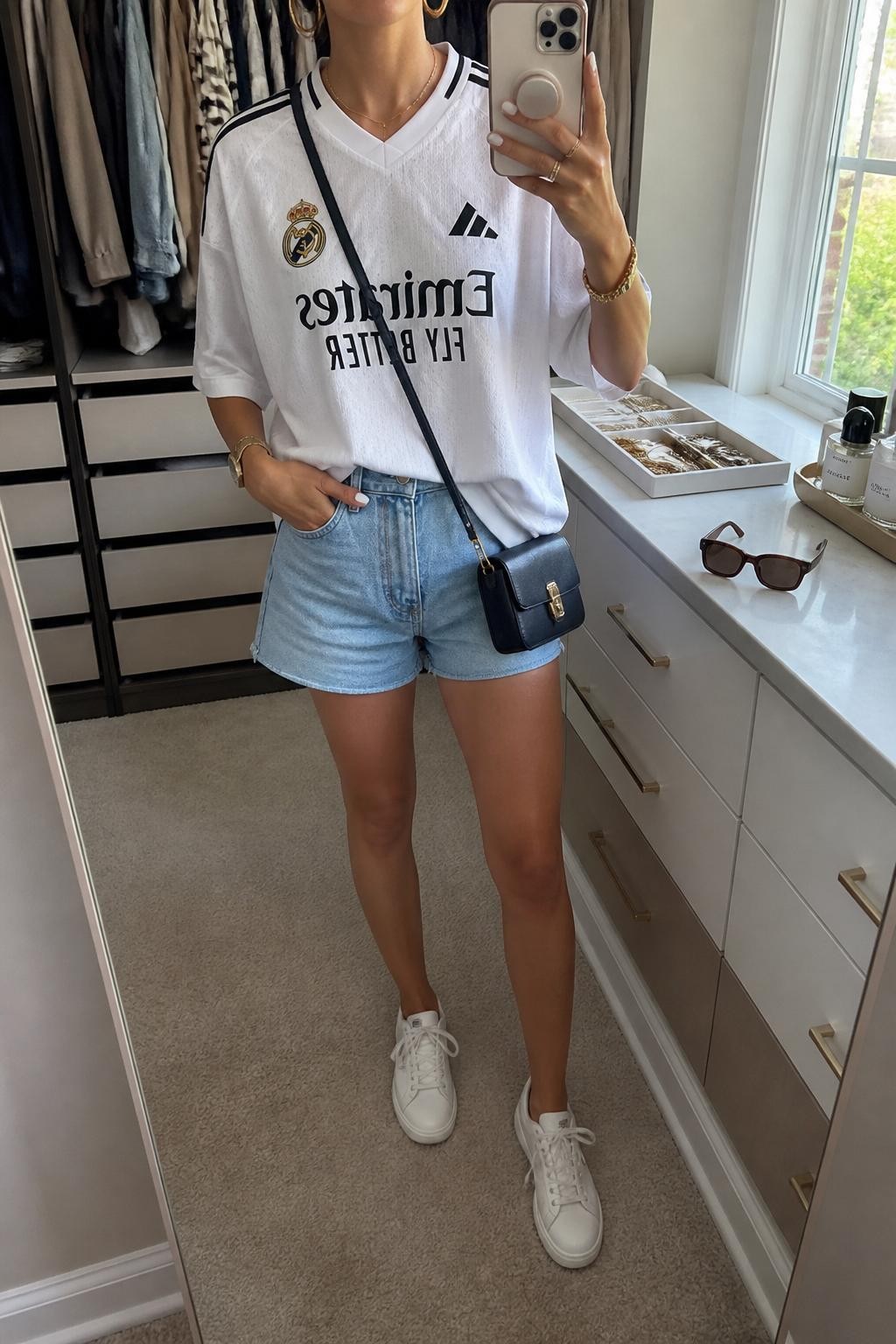 mirror selfie of a woman wearing an oversized white soccer jersey, light wash high-rise denim shorts, white low-top sneakers, navy mini crossbody bag, and gold hoop earrings, clearly showing the same clothing items, colors, fabrics, and accessories listed in the Outfit Pieces section, holding a phone in front of a mirror, cropped at NECK so the face does not show, with a relaxed casual selfie pose, in a walk-in closet mirror, natural window light, including sunglasses placed on a dresser and a jewelry tray on the vanity, latest iPhone photo quality.