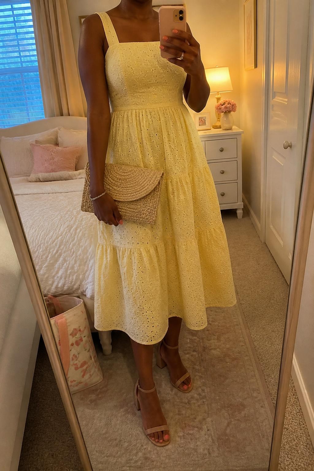 mirror selfie of a black woman wearing a butter yellow eyelet midi dress with nude block heel sandals, a natural woven clutch, pearl stud earrings, and rosy blush makeup, holding a phone in front of a mirror, cropped at NECK so face doesn't show, with a natural casual selfie pose, in a feminine-themed bedroom, evening golden hour, including small lived-in details like a tote bag on the floor and soft pillows on the bed, latest iPhone photo quality (no watermarks on images)