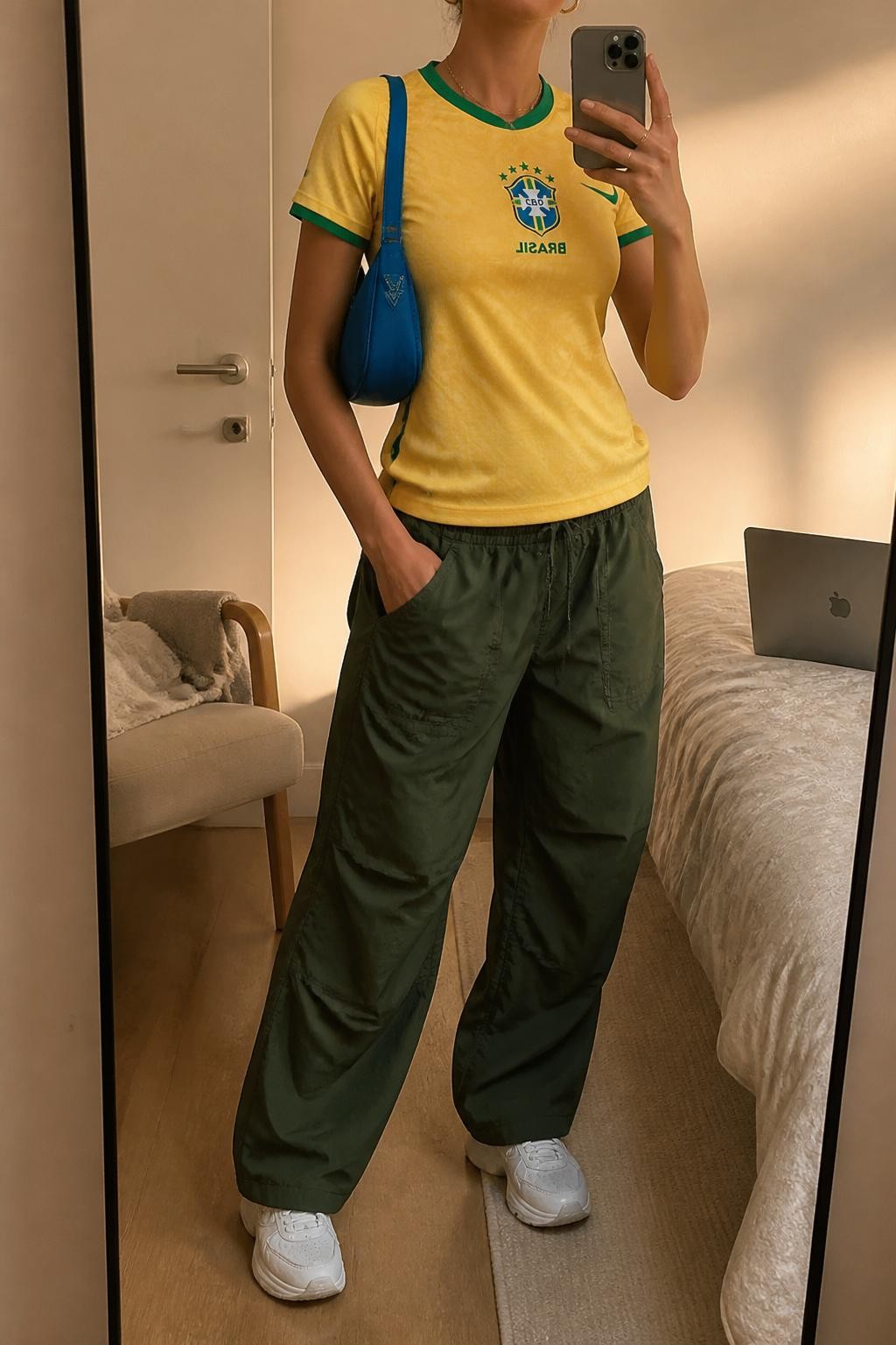 mirror selfie of a woman wearing a yellow Brazil soccer jersey, forest green parachute pants, white chunky sneakers, blue mini shoulder bag, and gold huggie earrings, clearly showing the same clothing items, colors, fabrics, and accessories listed in the Outfit Pieces section, holding a phone in front of a mirror, cropped at NECK so the face does not show, with a relaxed casual selfie pose, in a minimal dressing room, evening golden hour, including a folded blanket on a chair and a laptop on the bed, latest iPhone photo quality.