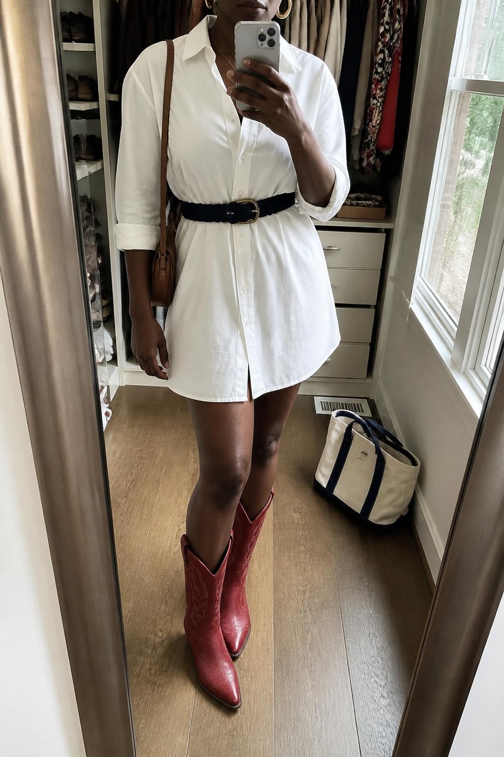 Outfit Image Prompt:
mirror selfie of a Black woman with rich dark skin wearing a white poplin button-front shirt dress, red western-style cowboy boots, a navy woven belt, a small tan shoulder bag, and gold hoop earrings, clearly showing the exact clothing items and accessories listed, holding a phone in front of a mirror, cropped at NECK so the face does not show, relaxed casual selfie pose, in a walk-in closet mirror, natural window light, with an open closet with clothes visible and a tote bag resting on the floor, realistic latest iPhone photo quality, natural skin texture.