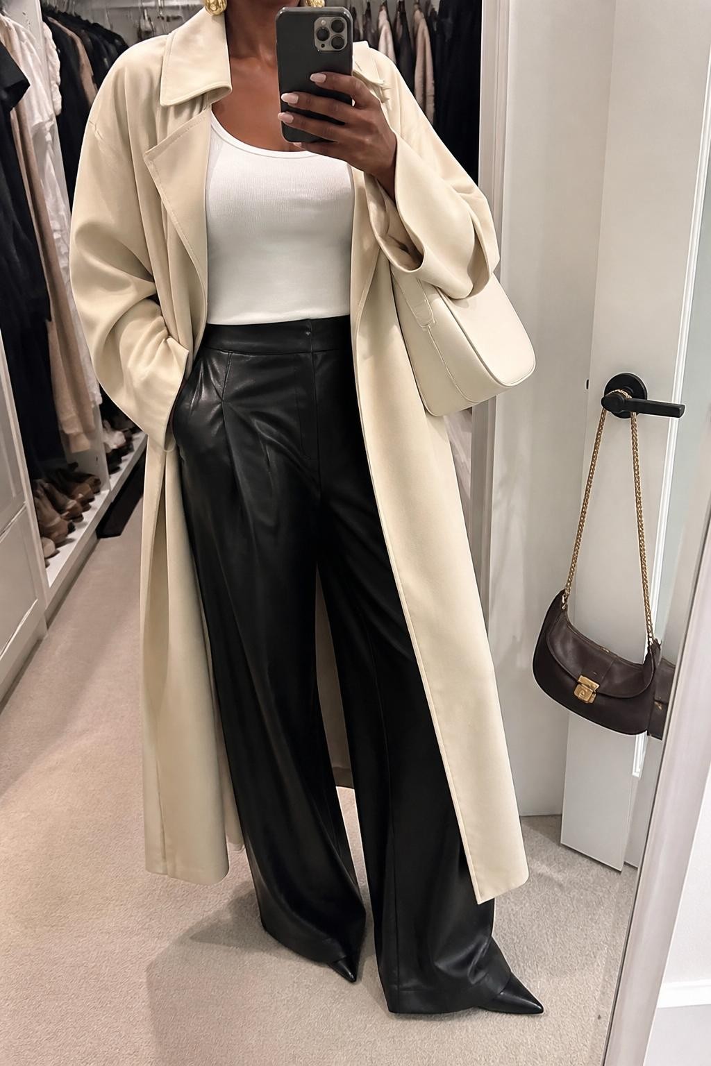 Outfit Image Prompt:
mirror selfie of a Black woman with rich deep brown skin wearing a cream longline trench coat, black wide-leg leather pants, fitted white tank top, pointed black ankle boots, structured cream shoulder bag, and gold sculptural earrings, holding a phone in front of a mirror, cropped at NECK so the face does not show, relaxed casual high-fashion selfie pose, in a walk-in closet mirror, soft indoor lighting, with an open closet with clothes visible and a handbag hanging on a door handle, natural skin texture, realistic latest iPhone photo quality.