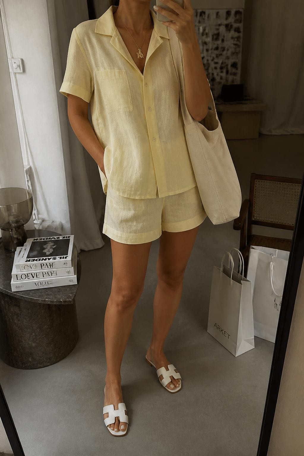 mirror selfie of a woman wearing a pale yellow linen short-sleeve button-up shirt, matching pale yellow linen shorts, white slide sandals, cream canvas tote bag, and gold charm necklace, clearly showing the same clothing items, colors, fabrics, and accessories listed in the Outfit Pieces section, holding a phone in front of a mirror, cropped at NECK so the face does not show, with a relaxed casual selfie pose, in a fashion studio mirror corner, warm moody lighting, including a stack of fashion magazines on a side table and shopping bags beside a chair, latest iPhone photo quality.