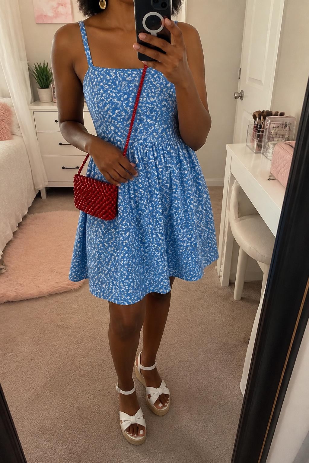 Outfit Image Prompt:
mirror selfie of a Black woman with warm dark brown skin wearing a blue floral cotton sundress, red beaded mini bag, white espadrille sandals, gold drop earrings, and a loose half-up natural hairstyle cropped at neck, clearly showing the exact clothing items and accessories listed, holding a phone in front of a mirror, cropped at NECK so the face does not show, relaxed casual selfie pose, in a feminine-themed bedroom, soft indoor lighting, with a small plant on a dresser and a makeup brush bag on the vanity, realistic latest iPhone photo quality, natural skin texture.