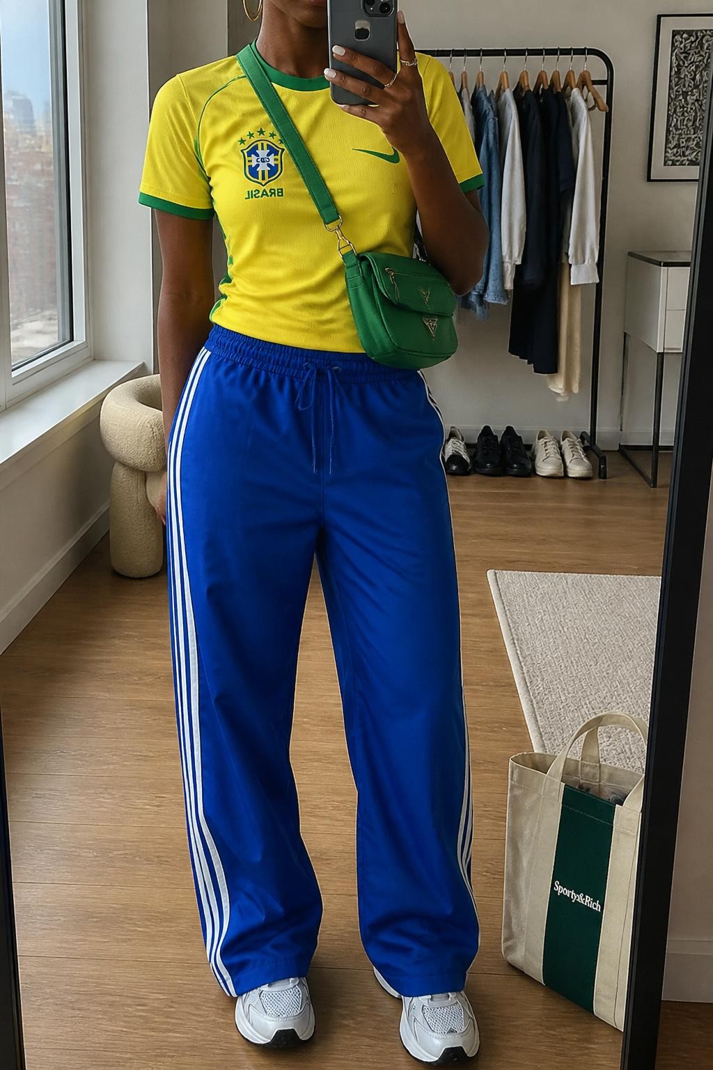 mirror selfie of a Black woman wearing a yellow Brazil soccer jersey, royal blue relaxed track pants with white side stripes, white running sneakers, green nylon crossbody bag, and silver hoop earrings, clearly showing the same clothing items, colors, fabrics, and accessories listed in the Outfit Pieces section, holding a phone in front of a mirror, cropped at NECK so the face does not show, with a relaxed casual selfie pose, in a stylish city apartment, natural window light, including a tote bag resting on the floor and clothing rack with garments behind, latest iPhone photo quality.