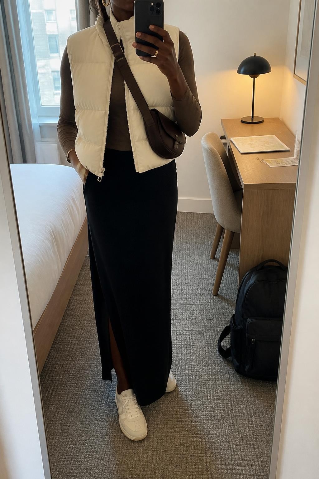 mirror selfie of a Black woman wearing the exact outfit described above, clearly showing an ivory puffer vest layered over a fitted mocha long-sleeve top, black column maxi skirt with a back slit, cream low-profile sneakers, a brown nylon crossbody bag, and gold hoop earrings, holding a phone in front of a mirror, cropped at NECK so the face does not show, with a relaxed casual selfie pose, in a clean hotel room mirror, soft morning light, with a backpack by the wall and a folded map or train pass on the desk, latest iPhone photo quality.