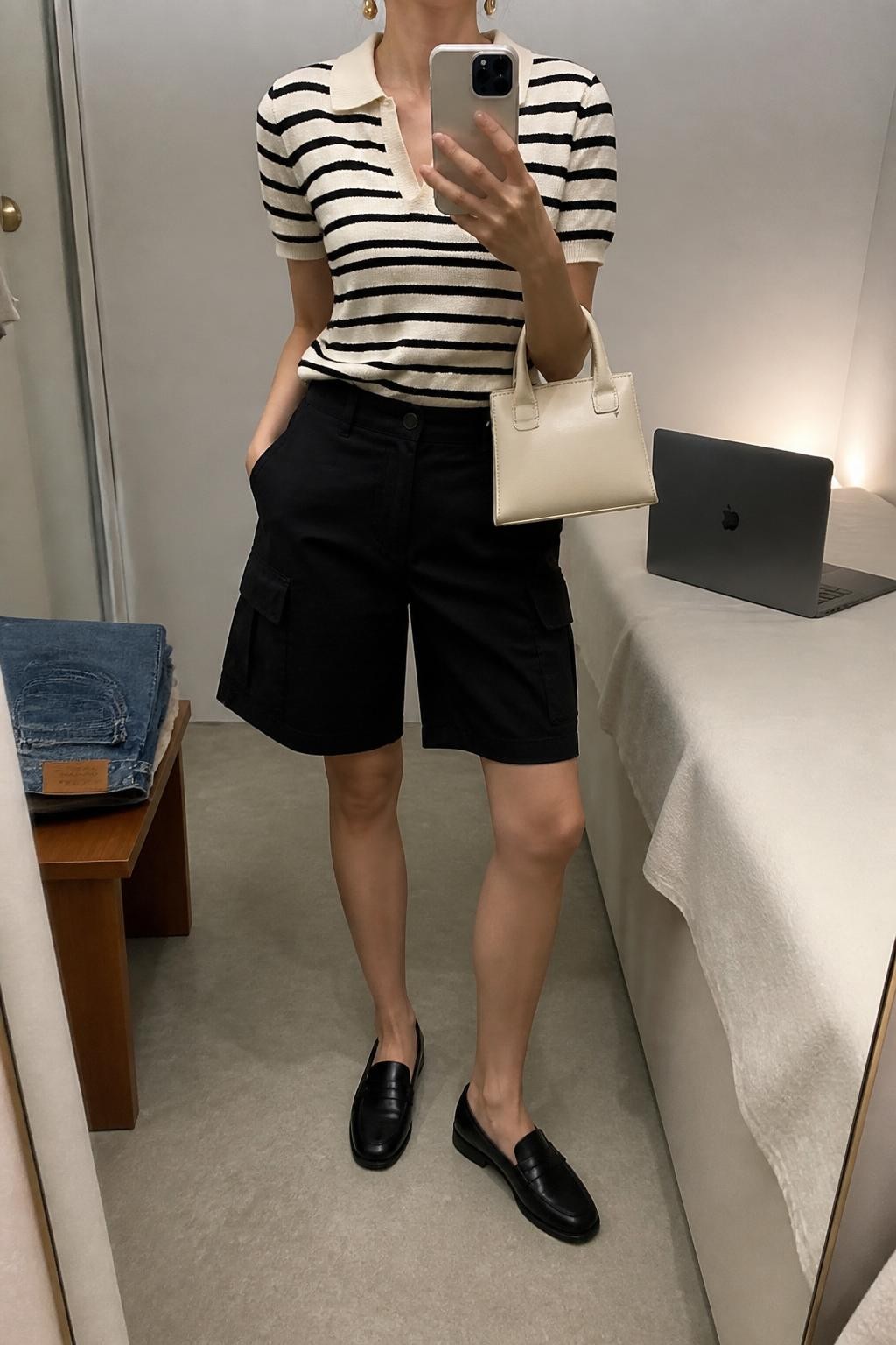 mirror selfie of a woman wearing black baggy cargo shorts, cream and black striped short-sleeve knit polo, black loafers, cream structured mini bag, and gold drop earrings, clearly showing the same clothing items, colors, fabrics, and accessories listed in the Outfit Pieces section, holding a phone in front of a mirror, cropped at NECK so the face does not show, with a relaxed casual selfie pose, in a minimal dressing room, soft indoor lighting, including a laptop on the bed and folded denim jeans on a bench, latest iPhone photo quality.