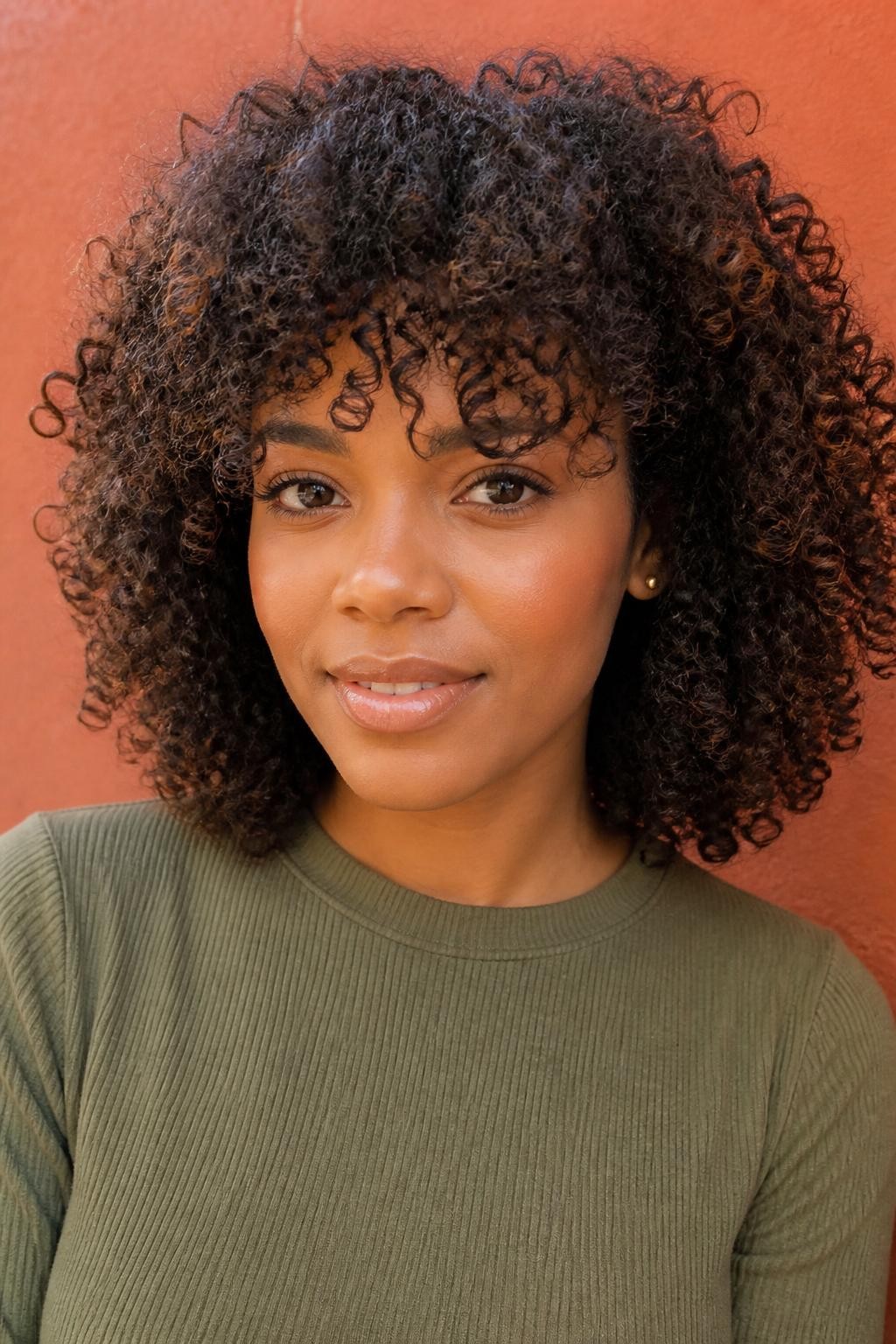A realistic close-up portrait of a Black woman with warm medium-dark skin wearing rounded curly hair with messy textured curl fringe, dark chocolate brown curls with subtle caramel tips on a few front curls, fringe made of soft uneven curls falling around the brows and upper forehead, longer curls around the temples, rounded shoulder-length shape, natural volume, defined curls with soft frizz, natural baby hairs, gold studs, olive green ribbed top, photographed against a warm terracotta wall in natural daylight, realistic curl pattern, natural skin detail, editorial beauty portrait, no overly perfect curl uniformity.