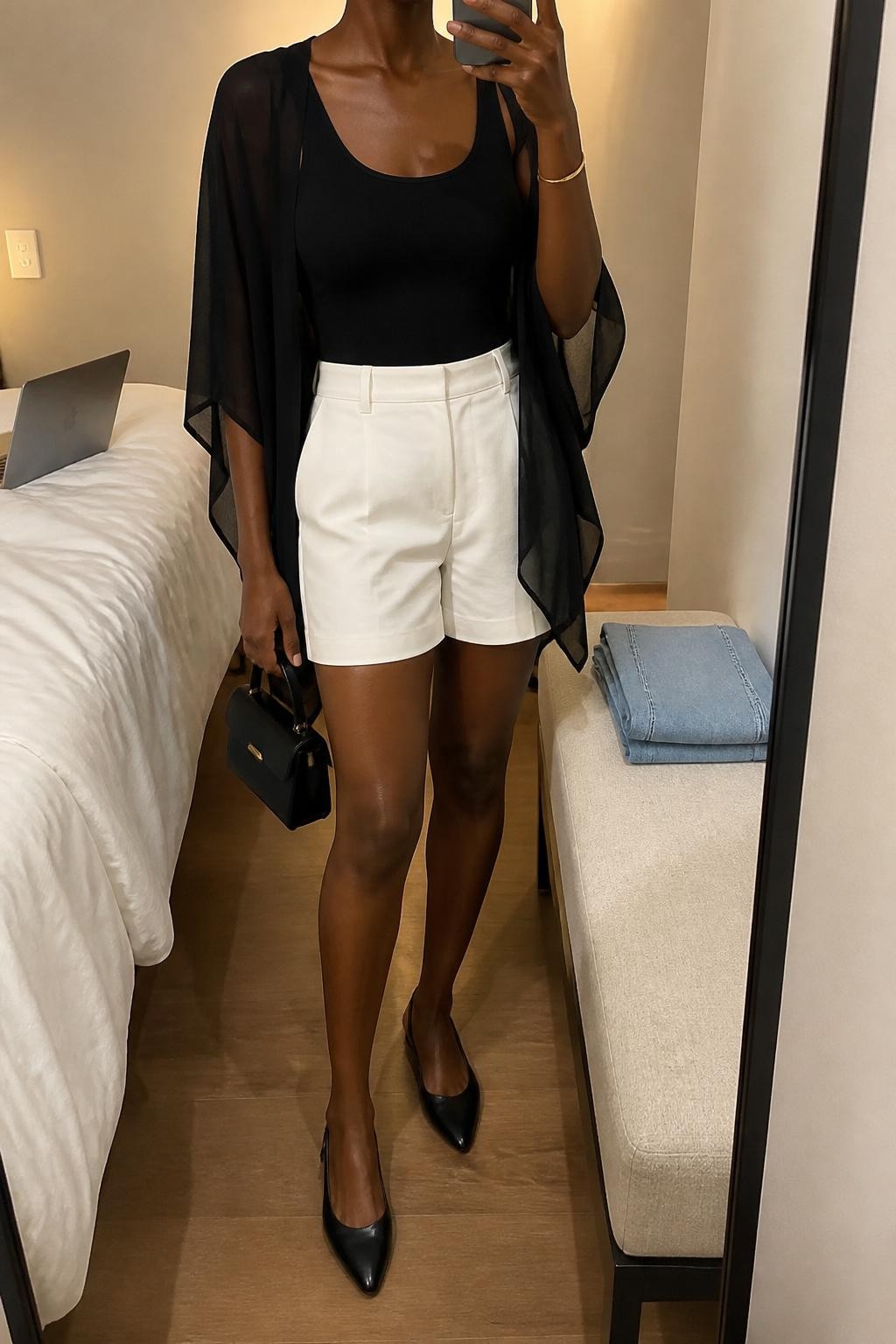 mirror selfie of a Black woman wearing a black sheer poncho layered over a black fitted tank top, white tailored high-waisted shorts, black slingback flats, black structured mini bag, and slim gold bangle bracelet, clearly showing the same clothing items, colors, fabrics, and accessories listed in the Outfit Pieces section, holding a phone in front of a mirror, cropped at NECK so the face does not show, with a relaxed casual selfie pose, in a minimal dressing room, evening golden hour, including a laptop on the bed and folded denim jeans on a bench, latest iPhone photo quality.