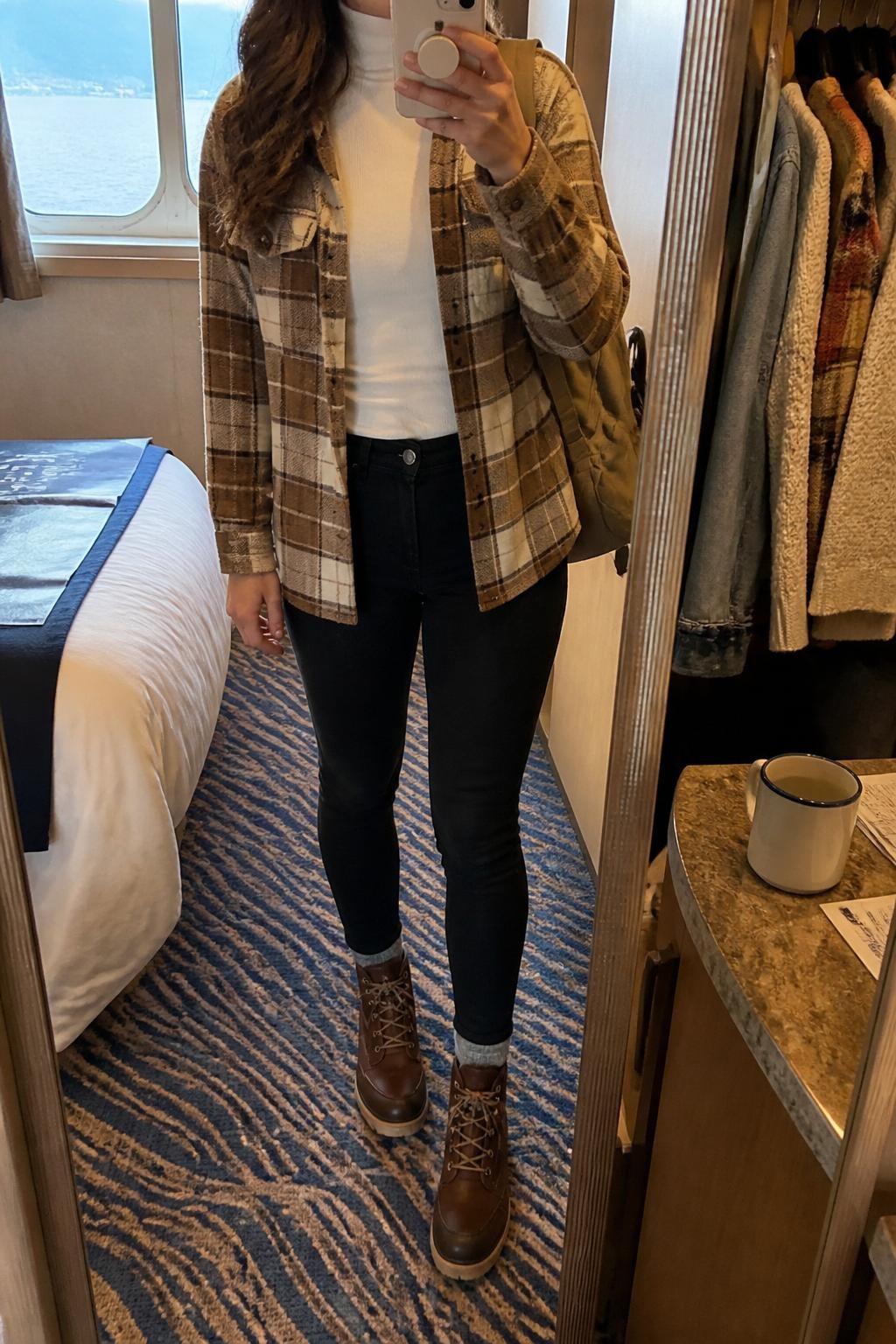 Outfit Image Prompt:
mirror selfie of a woman wearing a white fitted turtleneck, brown and cream plaid shacket, black skinny jeans, brown lace-up waterproof boots, a tan knit beanie, and a beige backpack, holding a phone in front of a mirror, cropped at NECK so the face does not show, relaxed casual selfie pose, in a cozy cruise cabin mirror setup with natural window light, including an open closet with clothes visible and a coffee mug on a small table, natural skin texture, realistic latest iPhone photo quality, casual travel outfit styling.