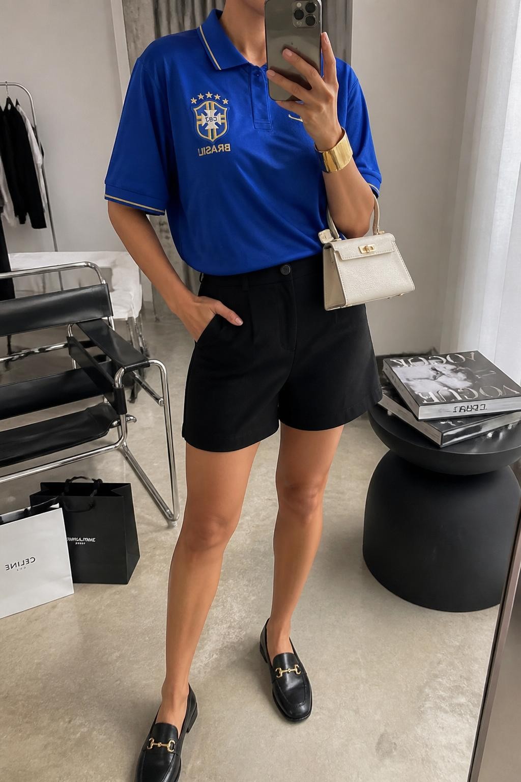 mirror selfie of a woman wearing an oversized royal blue soccer jersey tucked into black tailored high-waisted shorts, black loafers, cream structured mini bag, and gold cuff bracelet, clearly showing the same clothing items, colors, fabrics, and accessories listed in the Outfit Pieces section, holding a phone in front of a mirror, cropped at NECK so the face does not show, with a relaxed casual selfie pose, in a fashion studio mirror corner, soft indoor lighting, including a stack of fashion magazines on a side table and shopping bags beside a chair, latest iPhone photo quality.
