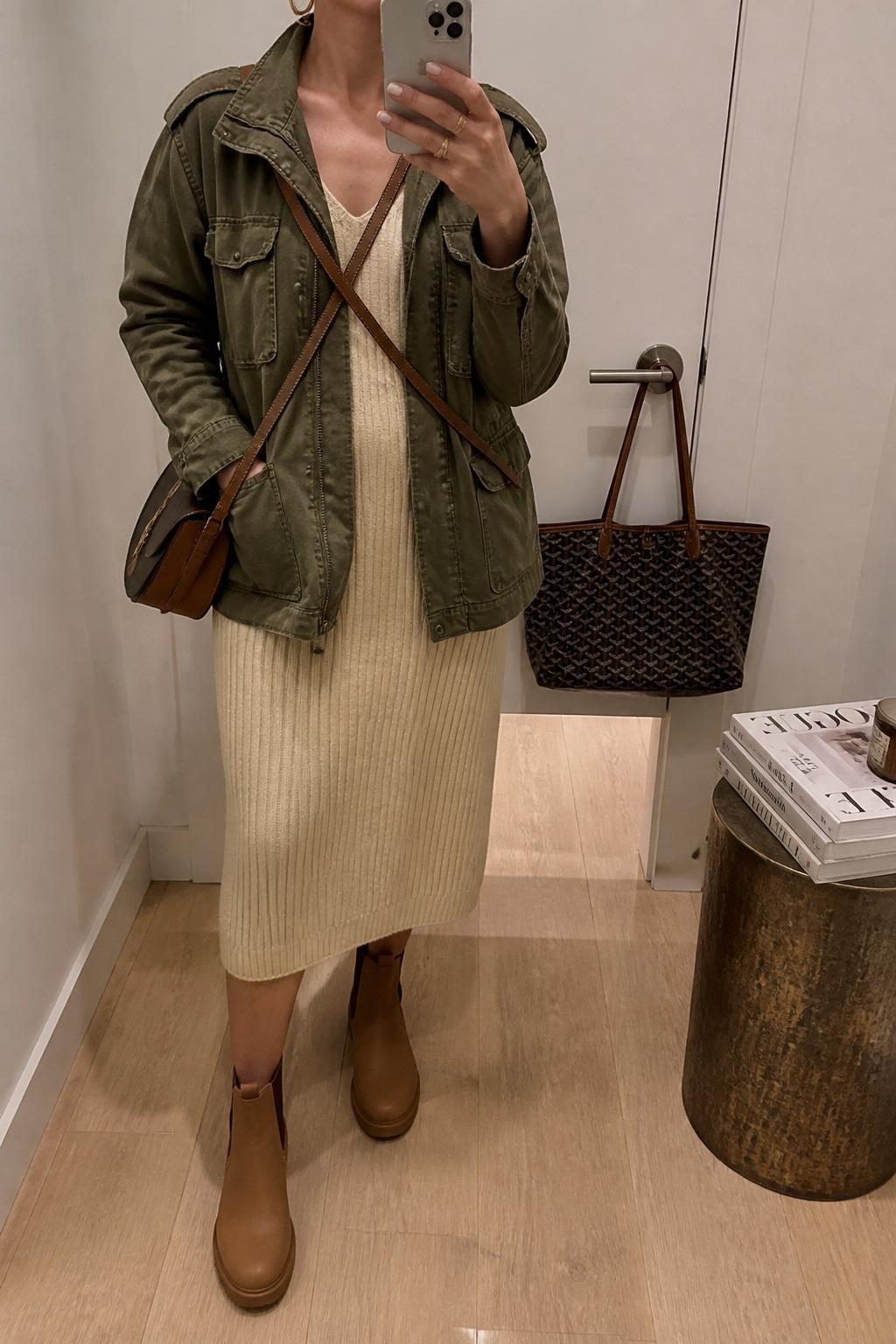 Outfit Image Prompt:
mirror selfie of a woman wearing an olive utility jacket, cream ribbed midi knit dress, tan waterproof ankle boots, a brown leather crossbody bag, and gold hoop earrings, holding a phone in front of a mirror, cropped at NECK so the face does not show, relaxed casual selfie pose, in a minimal dressing room with soft indoor lighting, including a handbag hanging on a door handle and a stack of fashion magazines on a side table, natural skin texture, latest iPhone photo quality, realistic casual mirror selfie.
