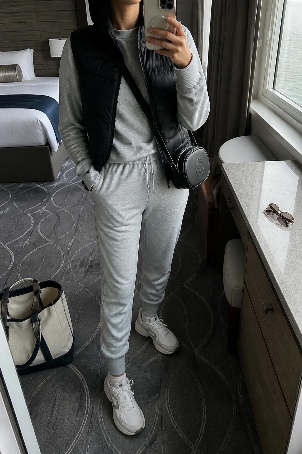 Outfit Image Prompt:
mirror selfie of a woman wearing a black lightweight puffer vest, gray matching jogger set with a soft crewneck sweatshirt, white chunky sneakers, a black beanie, and a small black crossbody bag, holding a phone in front of a mirror, cropped at NECK so the face does not show, relaxed casual selfie pose, in a stylish cruise cabin mirror corner with natural window light, including sunglasses placed on a dresser and a tote bag resting on the floor, realistic latest iPhone photo quality, natural skin texture, casual not staged.