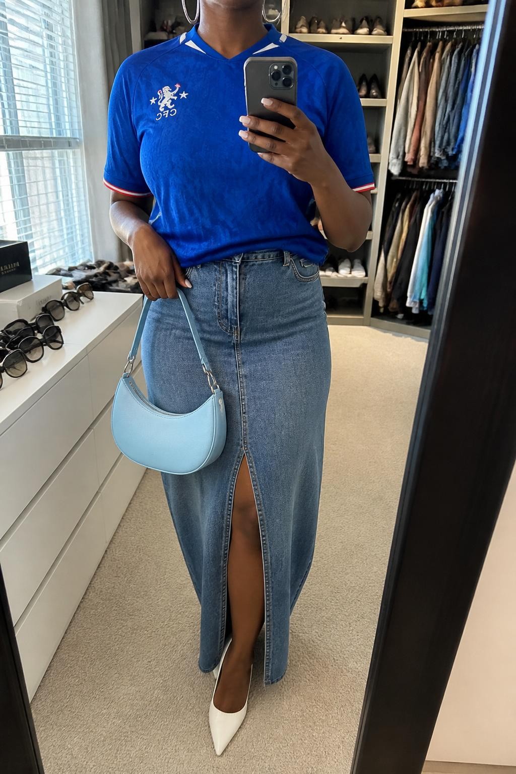 mirror selfie of a Black woman wearing a royal blue soccer jersey, medium blue denim maxi skirt with front slit, white pointed heels, pale blue crescent shoulder bag, and silver hoop earrings, clearly showing the same clothing items, colors, fabrics, and accessories listed in the Outfit Pieces section, holding a phone in front of a mirror, cropped at NECK so the face does not show, with a relaxed casual selfie pose, in a walk-in closet mirror, natural window light, including an open closet with clothes visible and sunglasses placed on a dresser, latest iPhone photo quality.