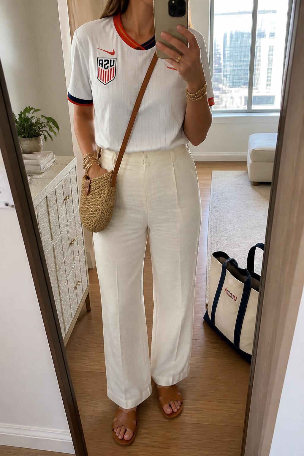 mirror selfie of a woman wearing a white USA soccer jersey with red and navy accents, cream high-waisted linen pants, tan leather slide sandals, woven straw crossbody bag, and gold layered bracelets, clearly showing the same clothing items, colors, fabrics, and accessories listed in the Outfit Pieces section, holding a phone in front of a mirror, cropped at NECK so the face does not show, with a relaxed casual selfie pose, in a stylish city apartment, soft indoor lighting, including a tote bag resting on the floor and a small plant on a dresser, latest iPhone photo quality.