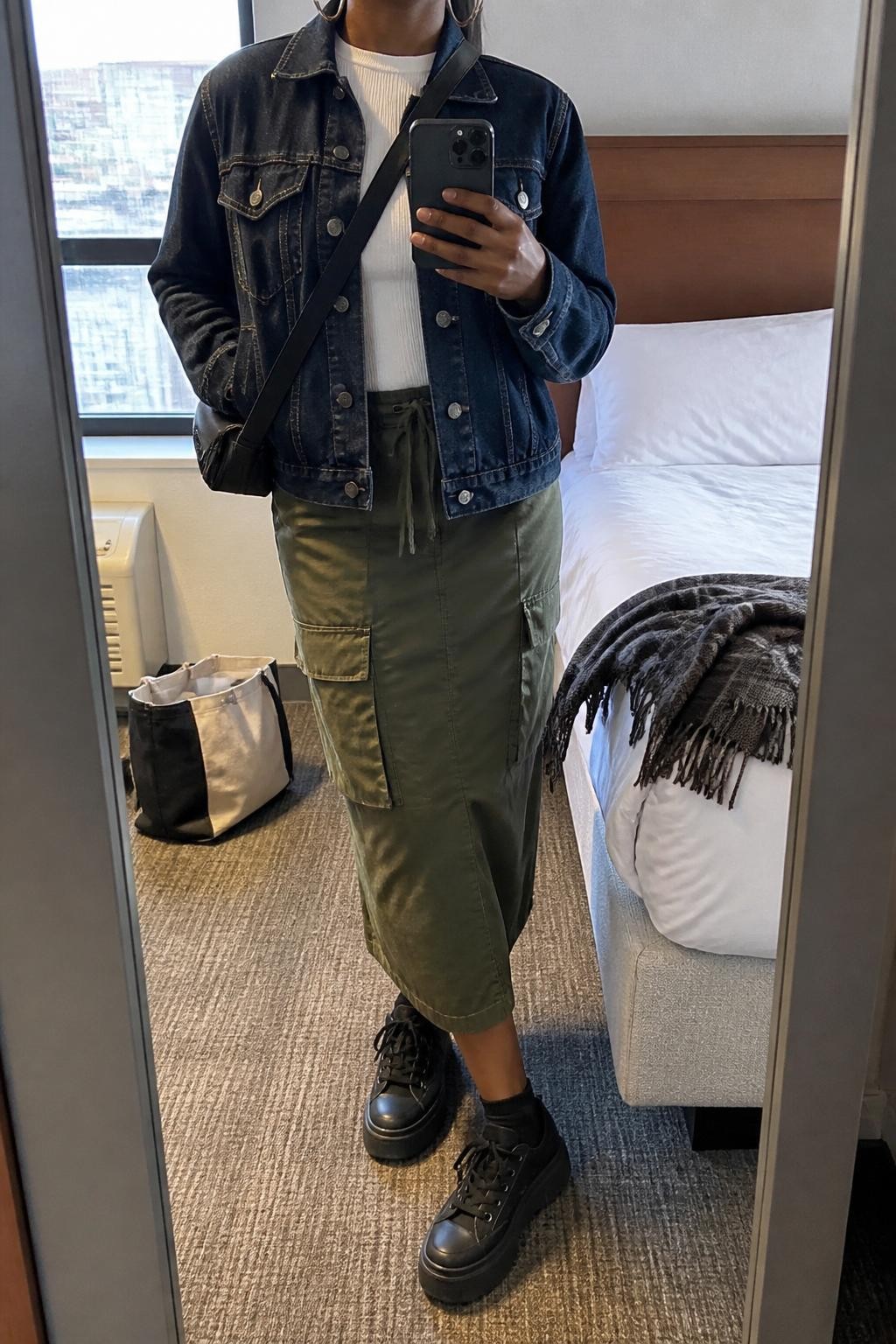 mirror selfie of a Black woman wearing the exact outfit described above, clearly showing a dark denim jacket layered over a fitted white ribbed top, olive cargo midi skirt, black platform sneakers, a black crossbody bag, and silver hoop earrings, holding a phone in front of a mirror, cropped at NECK so the face does not show, with a relaxed casual selfie pose, in a casual hotel room mirror, natural window light, with a tote bag resting on the floor and a folded scarf on the bed, latest iPhone photo quality.