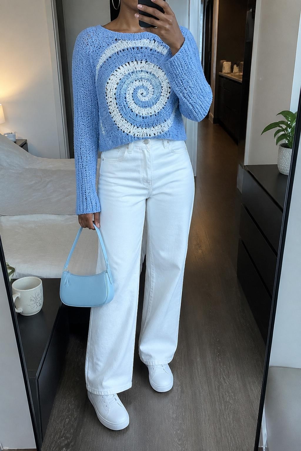 mirror selfie of a Black woman wearing a sky blue spiral crochet sweater with white swirl stitching, white high-waisted wide-leg jeans, white low-top sneakers, pale blue mini handbag, and silver hoop earrings, clearly showing the same clothing items, colors, fabrics, and accessories listed in the Outfit Pieces section, holding a phone in front of a mirror, cropped at NECK so the face does not show, with a relaxed casual selfie pose, in a stylish city apartment, soft indoor lighting, including a coffee mug on a bedside table and a small plant on a dresser, latest iPhone photo quality.