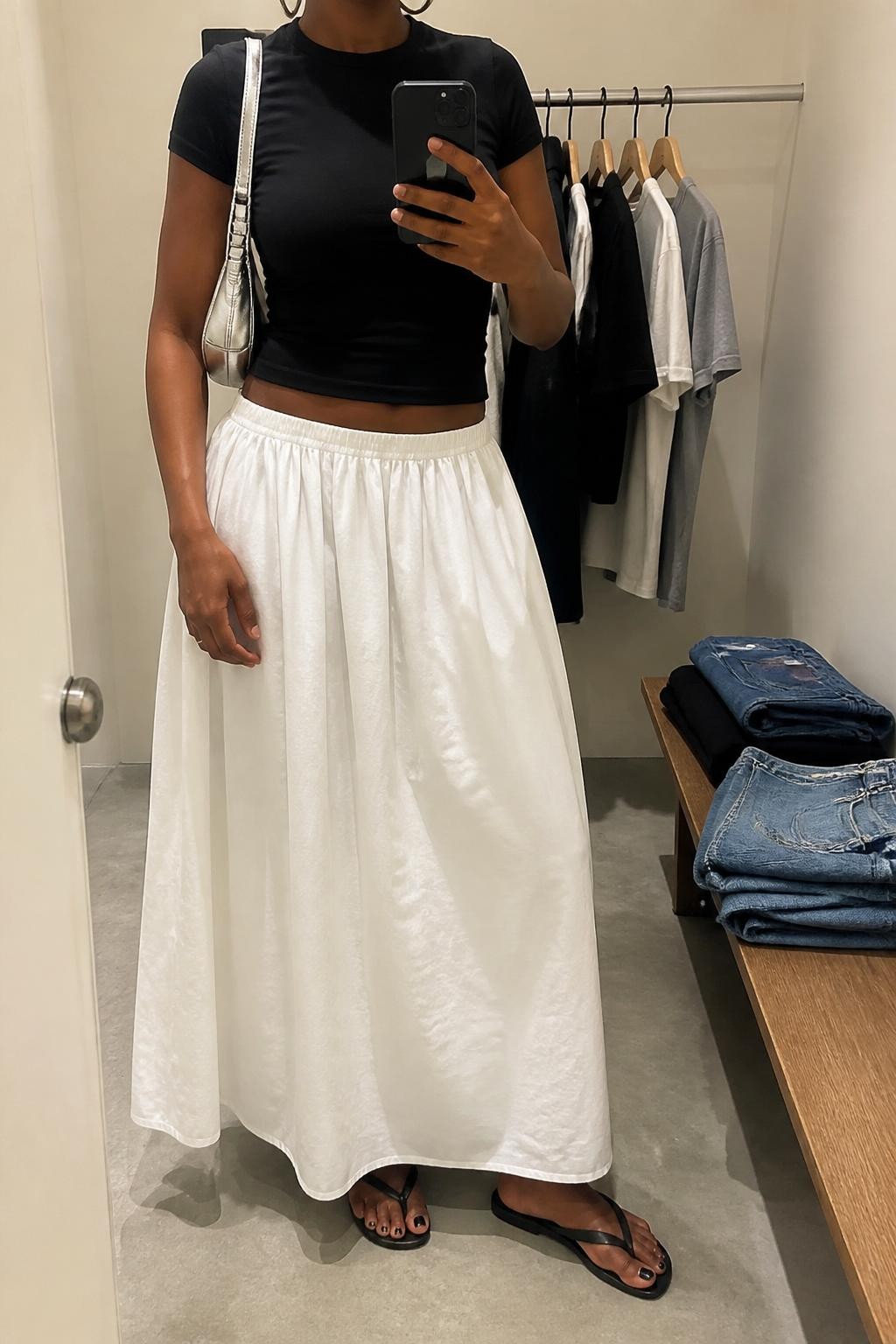 Outfit Image Prompt:
mirror selfie of a Black woman with rich brown skin wearing a fitted black baby tee, white cotton poplin maxi skirt, black slim flip-flop sandals, small silver shoulder bag, and silver hoop earrings, holding a phone in front of a mirror, cropped at NECK so the face does not show, relaxed casual selfie pose, in a minimal dressing room, soft indoor lighting, with a clothing rack with garments behind and folded denim jeans on a bench, realistic latest iPhone photo quality, natural skin texture.