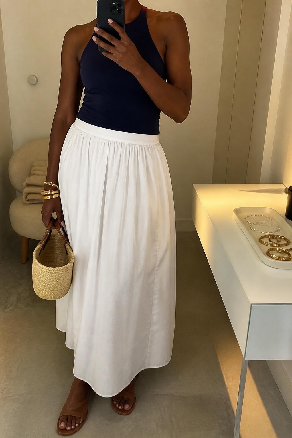 Outfit Image Prompt:
mirror selfie of a Black woman with deep brown skin wearing a navy fitted halter top, white cotton poplin maxi skirt, tan flat sandals, a small woven basket bag, and gold bangle bracelets, holding a phone in front of a mirror, cropped at NECK so the face does not show, relaxed casual selfie pose, in a minimal dressing room, evening golden hour lighting, with a folded blanket on a chair and a jewelry tray on the vanity, realistic latest iPhone photo quality, natural skin texture, soft summer styling.