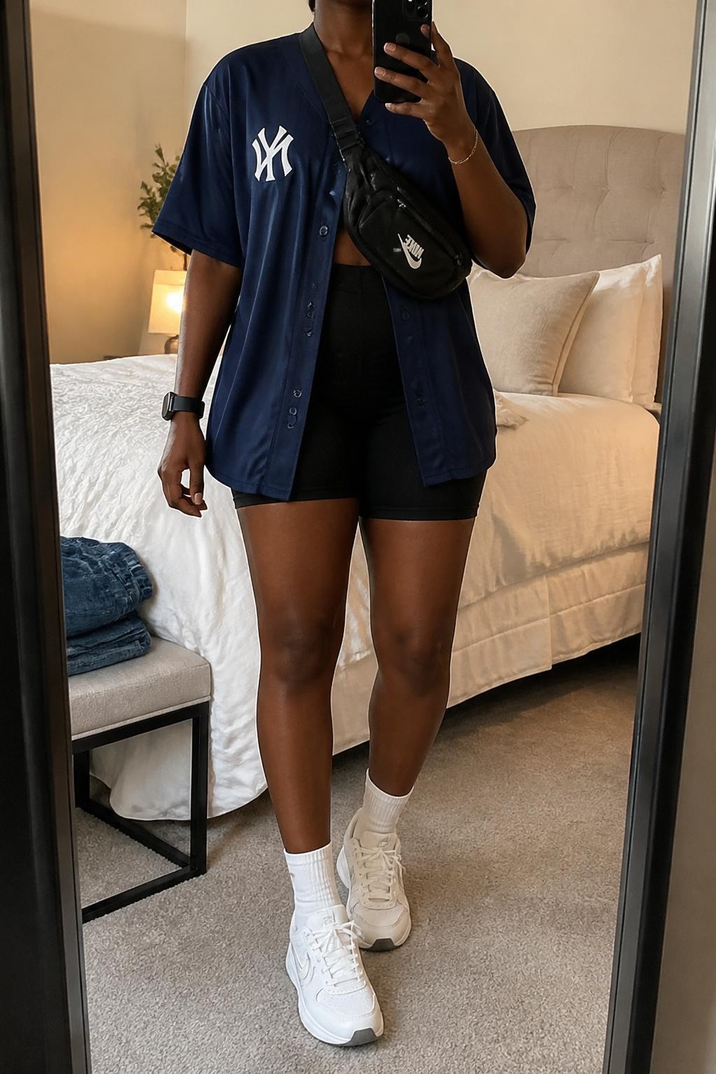 mirror selfie of a Black woman wearing an oversized navy button-front baseball jersey, black high-waisted biker shorts, white athletic sneakers, black nylon belt bag worn crossbody, and white crew socks, clearly showing the same clothing items, colors, fabrics, and accessories listed in the Outfit Pieces section, holding a phone in front of a mirror, cropped at NECK so the face does not show, with a relaxed casual selfie pose, in a cozy bedroom mirror setup, soft indoor lighting, including soft pillows on an unmade bed and folded denim jeans on a bench, latest iPhone photo quality.