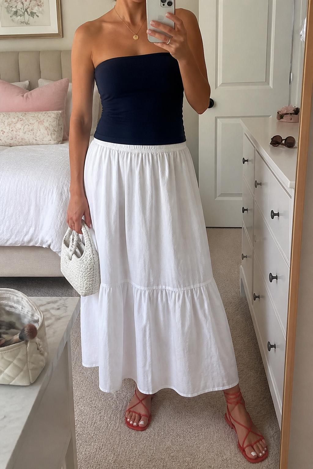 Image Prompt:
mirror selfie of a woman wearing a navy fitted tube top, white cotton poplin maxi skirt, red lace-up flat sandals, a small white woven handbag, and a gold pendant necklace, clearly showing the same clothing items, colors, fabrics, and accessories listed in the Outfit Pieces section, holding a phone in front of a mirror, cropped at NECK so the face does not show, with a relaxed casual selfie pose, in a feminine-themed bedroom, soft indoor lighting, with a makeup brush or cosmetic bag on the counter and sunglasses placed on a dresser, natural casual mirror selfie, not staged, latest iPhone photo quality.
