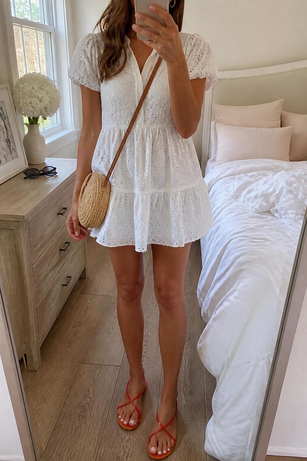 Image Prompt:
mirror selfie of a woman wearing a white eyelet mini dress with short sleeves, red strappy flat sandals, a small woven straw crossbody bag, dainty gold hoop earrings, and soft loose waves, clearly showing the same clothing items, colors, fabrics, and accessories listed in the Outfit Pieces section, holding a phone in front of a mirror, cropped at NECK so the face does not show, with a relaxed casual selfie pose, in a feminine-themed bedroom, natural window light, with sunglasses placed on a dresser and soft pillows on an unmade bed, natural casual mirror selfie, not staged, latest iPhone photo quality.