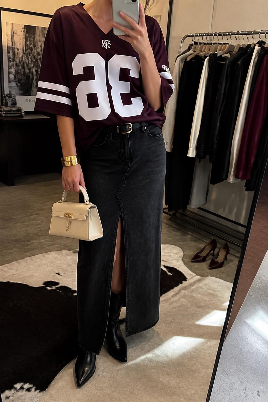 mirror selfie of a woman wearing an oversized burgundy football jersey, black denim maxi skirt with front slit, black pointed ankle boots, cream structured mini bag, and gold cuff bracelet, clearly showing the same clothing items, colors, fabrics, and accessories listed in the Outfit Pieces section, holding a phone in front of a mirror, cropped at NECK so the face does not show, with a relaxed casual selfie pose, in a fashion studio mirror corner, warm moody lighting, including a clothing rack with garments behind and a pair of heels on the floor nearby, latest iPhone photo quality.