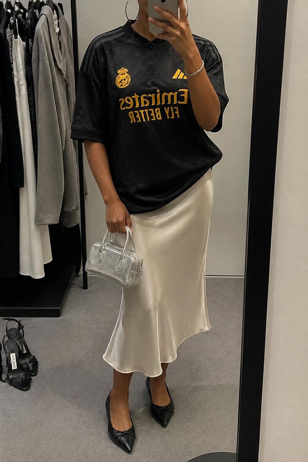 mirror selfie of a Black woman wearing an oversized black soccer jersey, ivory satin midi skirt, black pointed ballet flats, silver mini handbag, and chunky silver hoop earrings, clearly showing the same clothing items, colors, fabrics, and accessories listed in the Outfit Pieces section, holding a phone in front of a mirror, cropped at NECK so the face does not show, with a relaxed casual selfie pose, in a minimal dressing room, soft indoor lighting, including a clothing rack with garments behind and a pair of heels on the floor nearby, latest iPhone photo quality.
