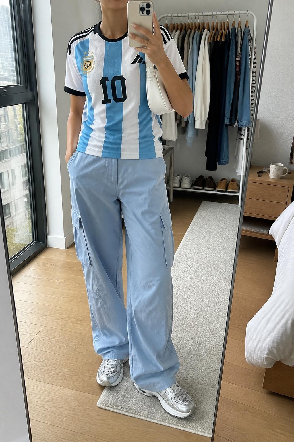 mirror selfie of a woman wearing a white Argentina soccer jersey, sky blue relaxed cargo pants, white and silver retro sneakers, white nylon shoulder bag, and small silver huggie earrings, clearly showing the same clothing items, colors, fabrics, and accessories listed in the Outfit Pieces section, holding a phone in front of a mirror, cropped at NECK so the face does not show, with a relaxed casual selfie pose, in a stylish city apartment, natural window light, including a clothing rack with garments behind and a coffee mug on a bedside table, latest iPhone photo quality.