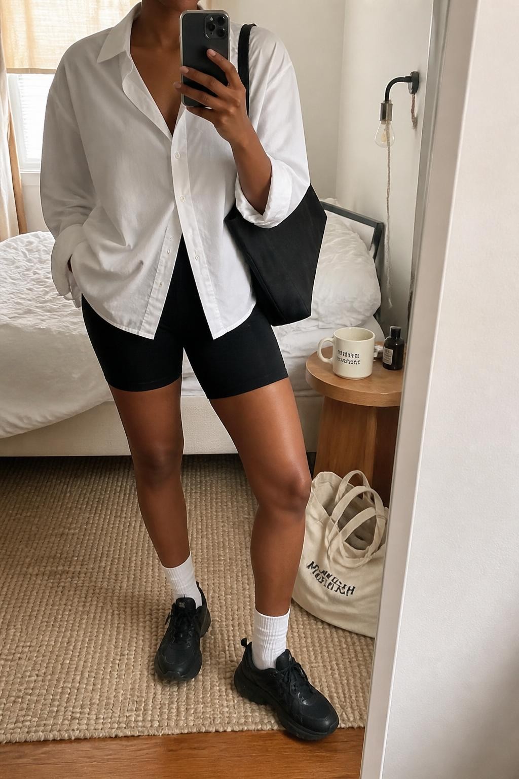 Outfit Image Prompt:
mirror selfie of a Black woman with medium brown skin wearing an oversized white poplin button-down shirt, black high-waisted bike shorts, white crew socks, black chunky sneakers, black rectangular sunglasses, and a black nylon tote bag, holding a phone in front of a mirror, cropped at NECK so the face does not show, relaxed casual NYC street style selfie pose, in a cozy bedroom mirror setup, natural window light, with a tote bag resting on the floor and a coffee mug on a bedside table, realistic latest iPhone photo quality, natural skin texture.