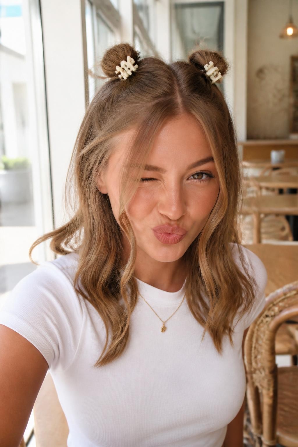 Image Prompt:
A realistic fashion beauty photo of a woman with medium-length dark blonde hair styled into two small half-up space buns secured with mini matte cream claw clips, loose waves left down, wispy face-framing strands, playful expression, white baby tee, bright café background, soft daylight, trendy youthful hairstyle inspiration, high-resolution DSLR photo, no text, (no watermarks on images)