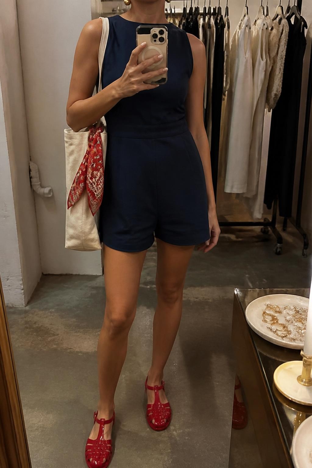 mirror selfie of a woman wearing a navy sleeveless linen romper, glossy red jelly shoes, white canvas tote bag, gold stud earrings, and a red silk scarf tied on the tote handle, clearly showing the same clothing items, colors, fabrics, and accessories listed in the Outfit Pieces section, holding a phone in front of a mirror, cropped at NECK so the face does not show, with a relaxed casual selfie pose, in a fashion studio mirror corner, warm moody lighting, including a clothing rack with garments behind and a jewelry tray on the vanity, latest iPhone photo quality.