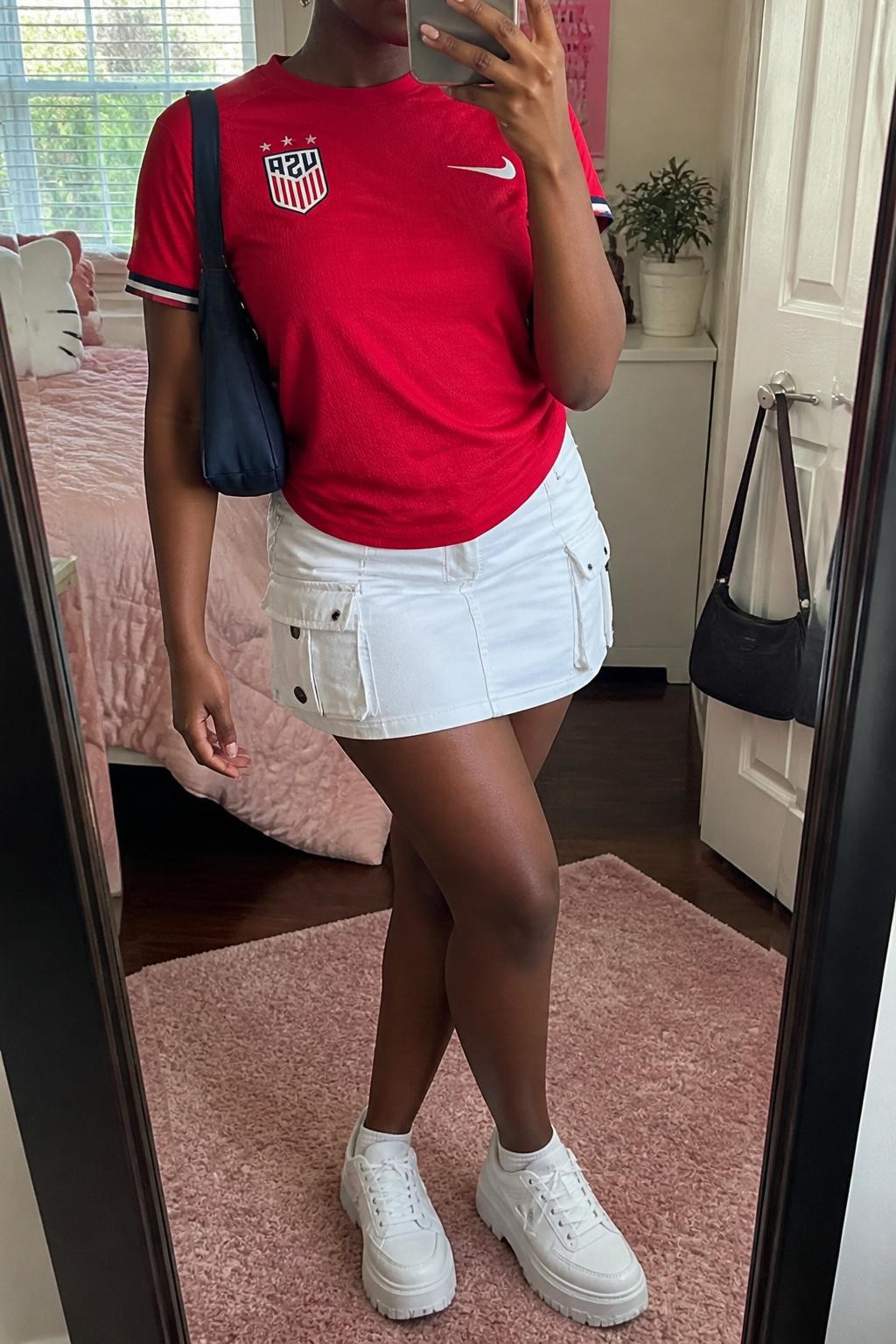mirror selfie of a Black woman wearing a red USA soccer jersey, white cargo mini skirt, white platform sneakers, navy nylon shoulder bag, and small silver huggie earrings, clearly showing the same clothing items, colors, fabrics, and accessories listed in the Outfit Pieces section, holding a phone in front of a mirror, cropped at NECK so the face does not show, with a relaxed casual selfie pose, in a feminine-themed bedroom, natural window light, including a handbag hanging on a door handle and a small plant on a dresser, latest iPhone photo quality.