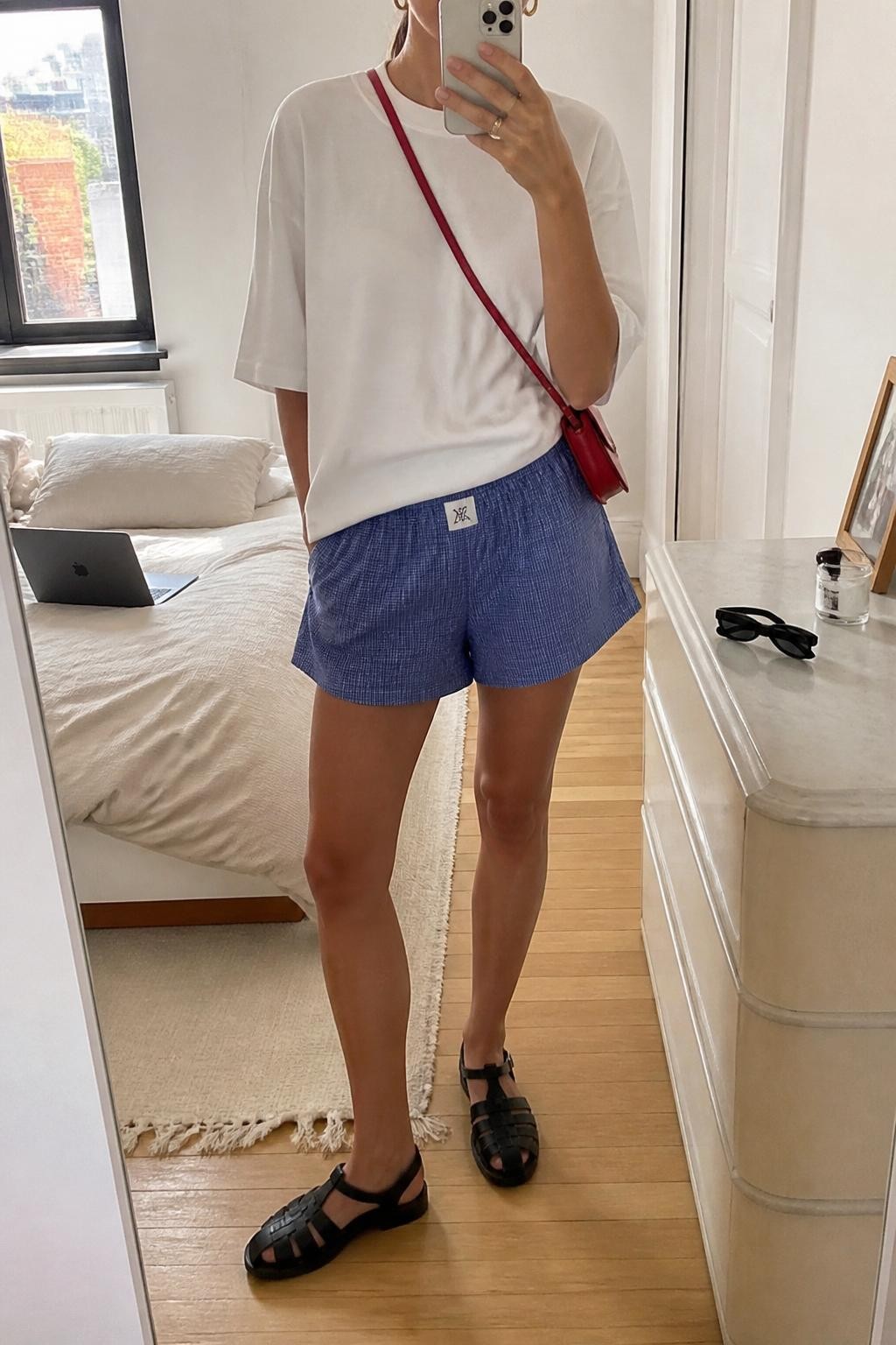 Outfit Image Prompt:
mirror selfie of a woman wearing an oversized white cotton tee, blue printed boxer-style shorts, black leather fisherman sandals, small red crossbody bag, and gold hoop earrings, holding a phone in front of a mirror, cropped at NECK so the face does not show, relaxed casual summer selfie pose, in a stylish city apartment, natural window light, with a laptop on the bed and sunglasses placed on a dresser, realistic latest iPhone photo quality, lived-in NYC outfit styling.