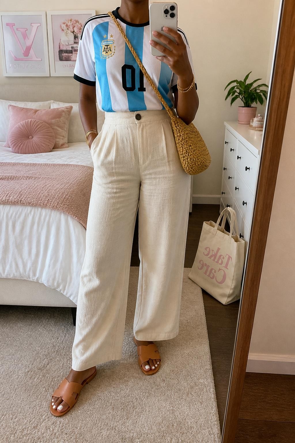 mirror selfie of a Black woman wearing a sky blue and white Argentina soccer jersey, cream high-waisted linen pants, tan leather slide sandals, woven straw crossbody bag, and gold layered bracelets, clearly showing the same clothing items, colors, fabrics, and accessories listed in the Outfit Pieces section, holding a phone in front of a mirror, cropped at NECK so the face does not show, with a relaxed casual selfie pose, in a feminine-themed bedroom, soft indoor lighting, including a small plant on a dresser and a tote bag resting on the floor, latest iPhone photo quality.