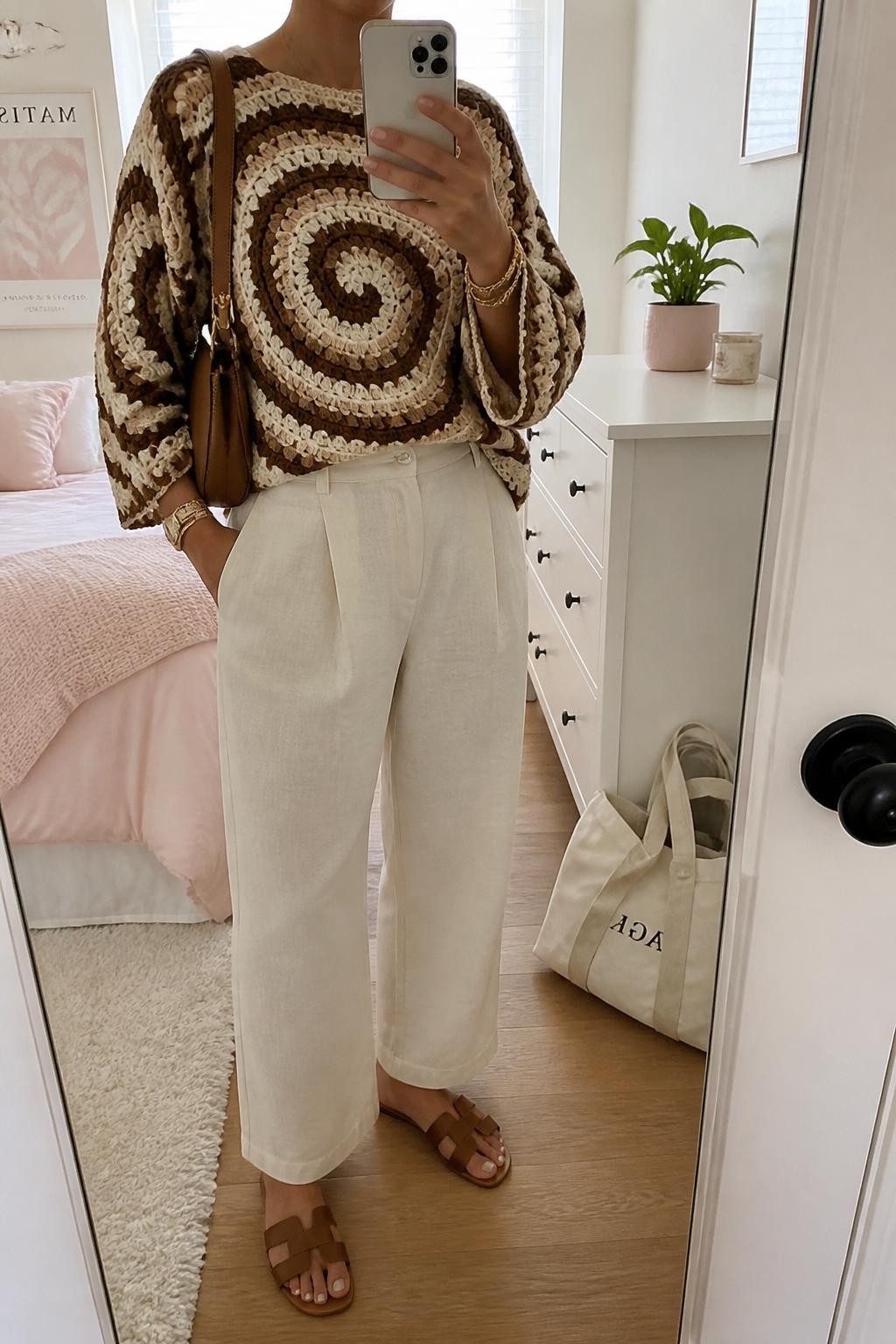 mirror selfie of a woman wearing a brown and cream spiral crochet sweater with textured circular crochet motifs, cream high-waisted linen pants, brown leather slide sandals, tan crescent shoulder bag, and gold layered bracelets, clearly showing the same clothing items, colors, fabrics, and accessories listed in the Outfit Pieces section, holding a phone in front of a mirror, cropped at NECK so the face does not show, with a relaxed casual selfie pose, in a feminine-themed bedroom, natural window light, including a small plant on a dresser and a cream tote bag resting on the floor, latest iPhone photo quality.