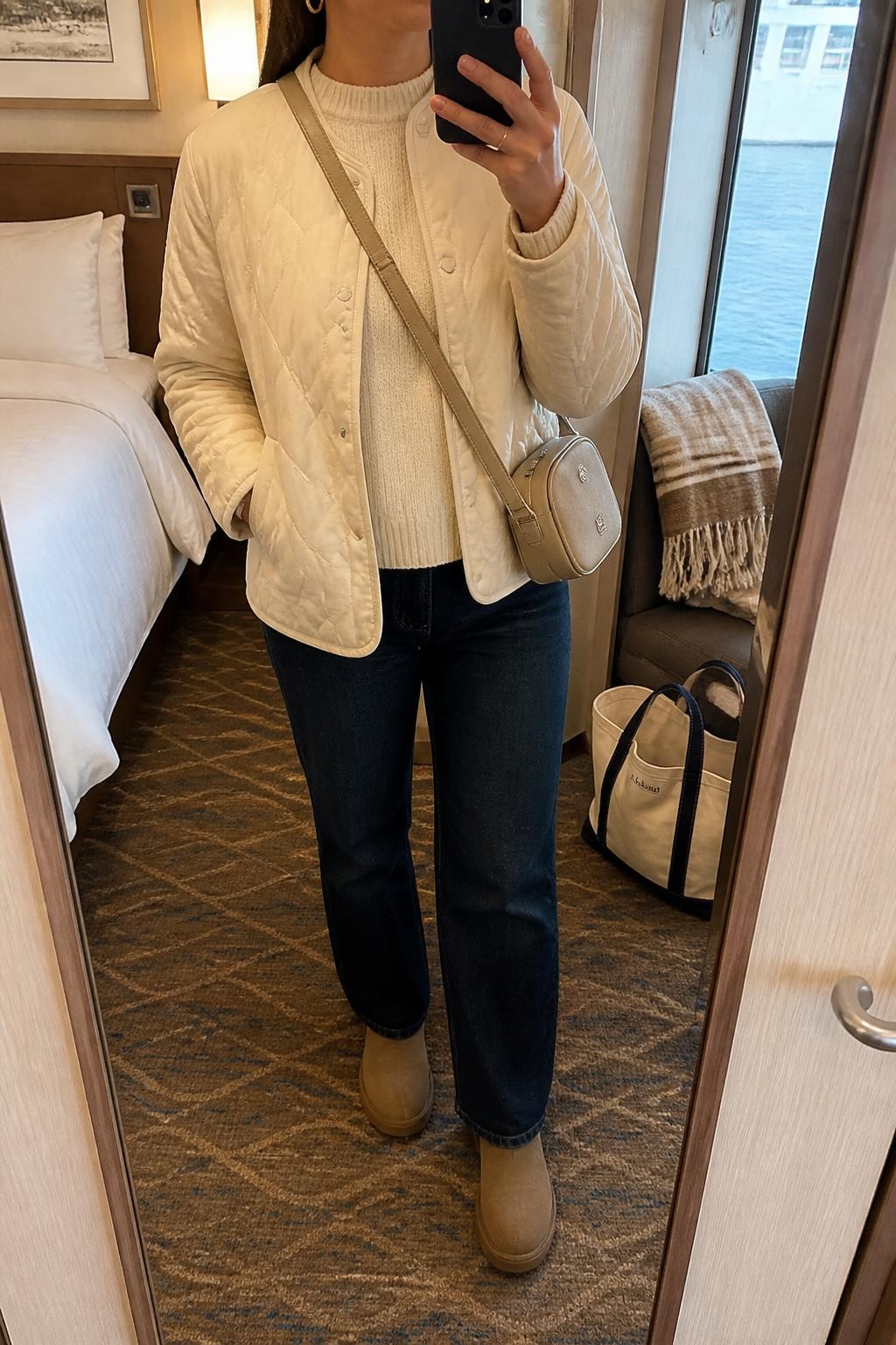 Outfit Image Prompt:
mirror selfie of a woman wearing a cream lightweight quilted jacket, ivory knit sweater, dark straight-leg jeans, tan waterproof ankle boots, a beige crossbody bag, and small gold hoop earrings, holding a phone in front of a mirror, cropped at NECK so the face does not show, relaxed casual selfie pose, in a cozy cruise cabin mirror setup with soft indoor lighting, including a folded blanket on a chair and a tote bag resting on the floor, natural skin texture, realistic latest iPhone photo quality, soft casual styling, not overly polished.