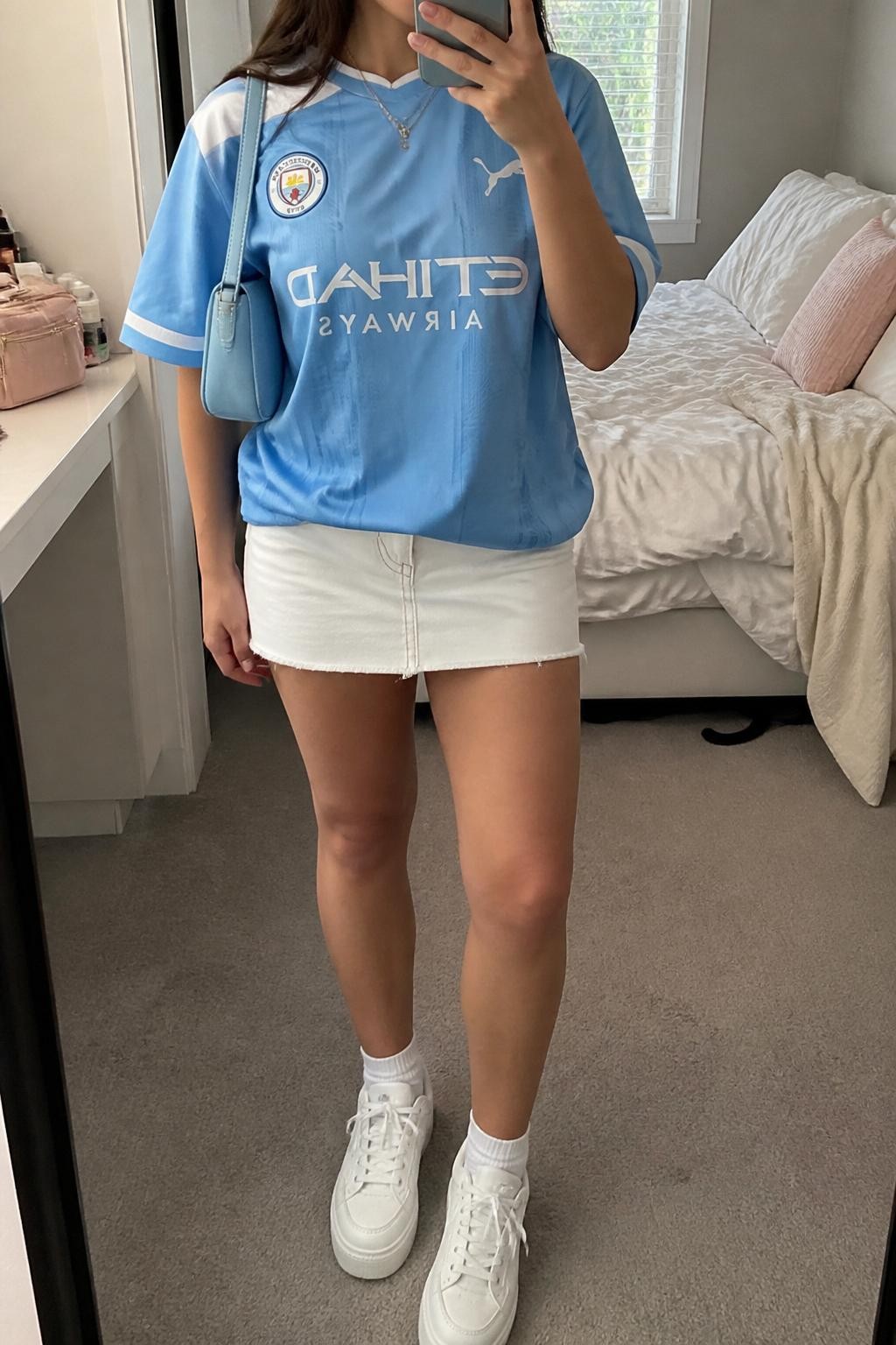 mirror selfie of a woman wearing an oversized sky blue soccer jersey, white denim mini skirt, white chunky sneakers, pale blue mini shoulder bag, and delicate silver necklace, clearly showing the same clothing items, colors, fabrics, and accessories listed in the Outfit Pieces section, holding a phone in front of a mirror, cropped at NECK so the face does not show, with a relaxed casual selfie pose, in a cozy bedroom mirror setup, natural window light, including soft pillows on an unmade bed and a cosmetic bag on the counter, latest iPhone photo quality.