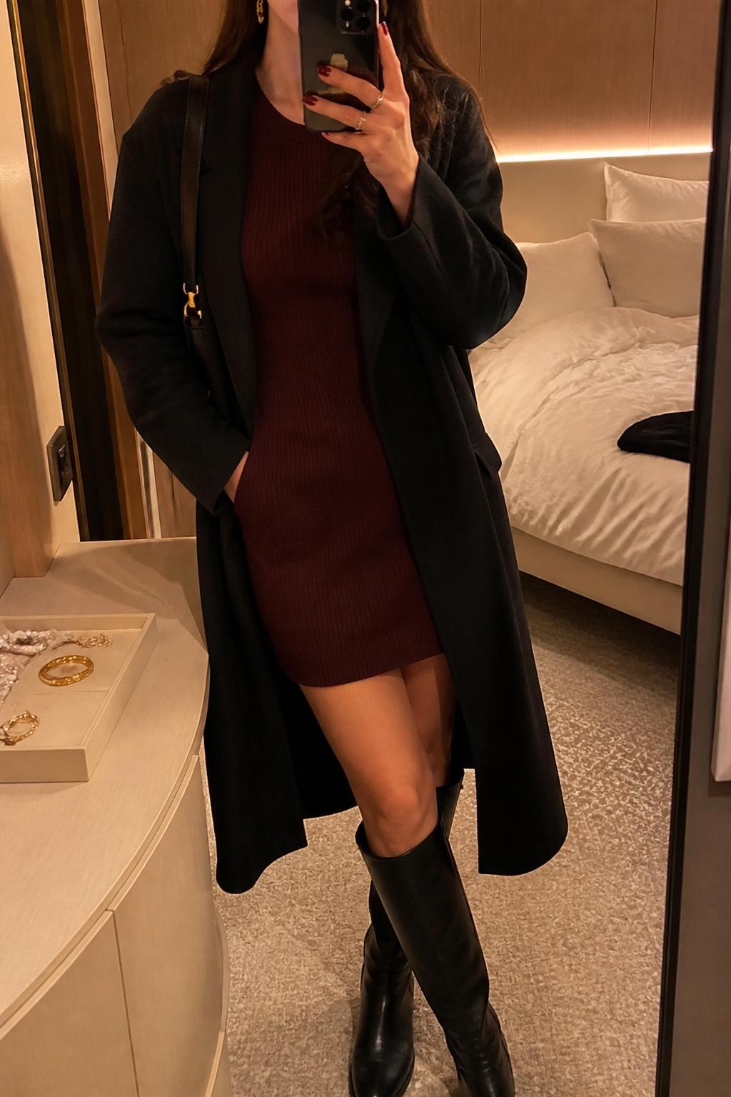 Outfit Image Prompt:
mirror selfie of a woman wearing a burgundy ribbed sweater dress, black tall boots, a black wool wrap coat, a small black shoulder bag, and gold drop earrings, holding a phone in front of a mirror, cropped at NECK so the face does not show, relaxed casual selfie pose, in a minimal cruise cabin mirror corner with warm moody lighting, including soft pillows on an unmade bed and a jewelry tray on the vanity, realistic latest iPhone photo quality, natural skin texture, soft dinner styling.
